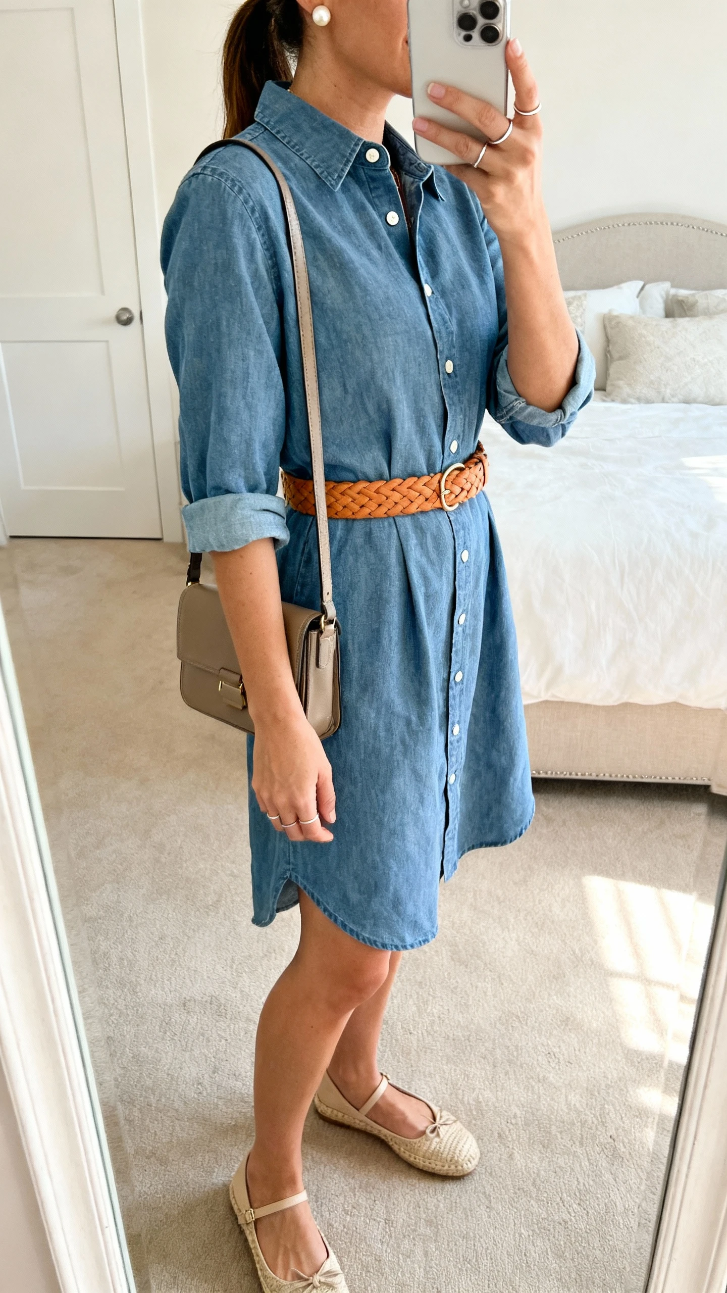 iPhone back/side-view selfie of a woman wearing a cotton or chambray shirt dress (collar, buttons to mid-torso, sleeves rolled) cinched with a tan or caramel woven belt, closed-toe espadrilles or classic ballet flats, a structured crossbody or satchel, pearl studs, and a slim ring stack; face not visible, bedroom mirror, soft afternoon light, iPhone photo quality.