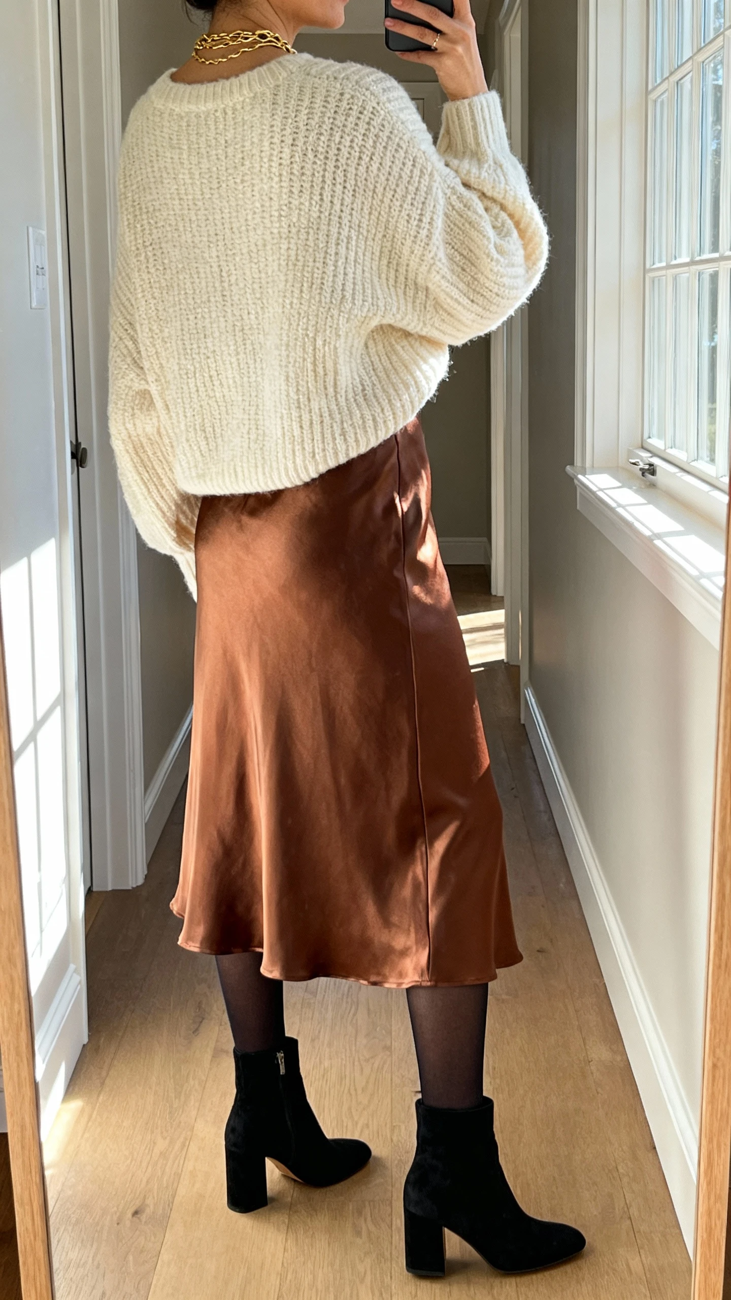 iPhone back-view hallway mirror selfie of a woman wearing a bronze satin midi skirt, chunky cream knit sweater (relaxed fit), black suede ankle boots with block heel, black opaque tights, layered gold necklaces, face not visible, natural daylight from window, iPhone photo quality.