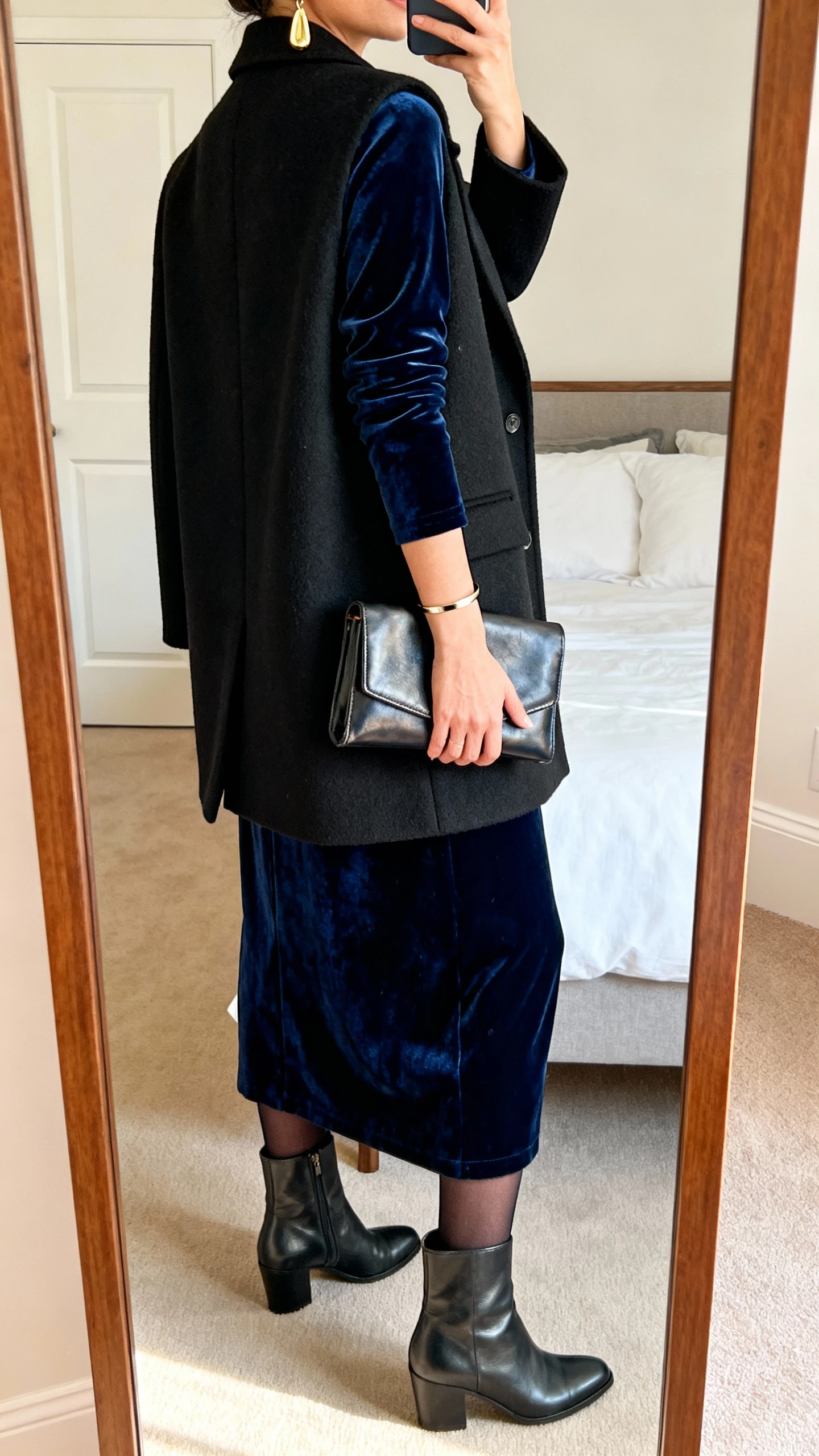 iPhone back-view mirror selfie of a woman in a midnight blue velvet long-sleeve maxi dress under a tailored black wool coat, sleek leather ankle boots, opaque tights, gold drop earrings, a slim cuff bracelet, and a leather clutch in hand; face not visible, bedroom mirror, warm indoor lighting, iPhone photo quality.