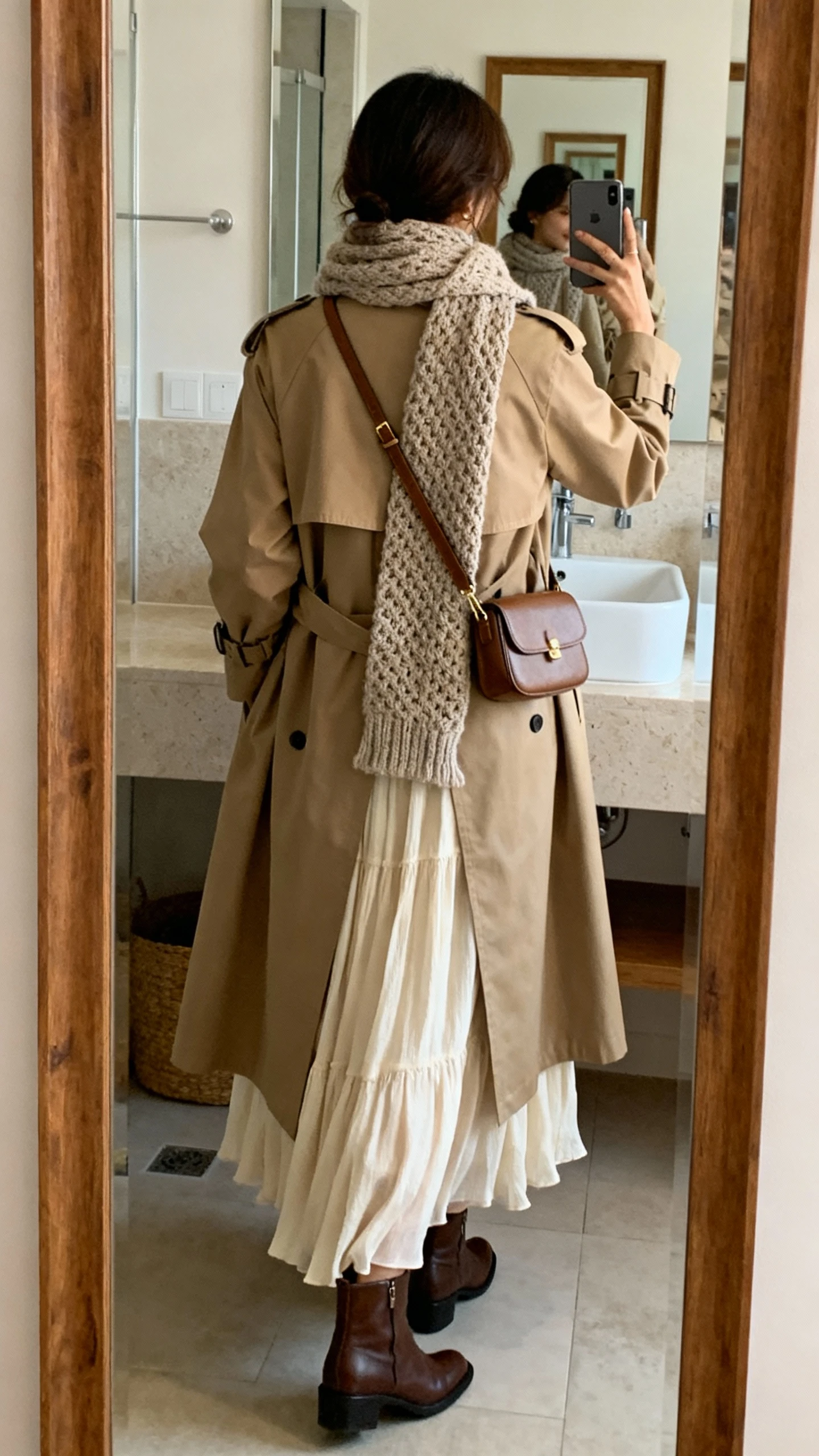 iPhone back-view mirror selfie of a woman wearing a flowy maxi dress layered under a classic trench coat, textured knit scarf, ankle boots, and a small crossbody bag, face not visible, bathroom mirror, indoor natural lighting, iPhone photo quality.