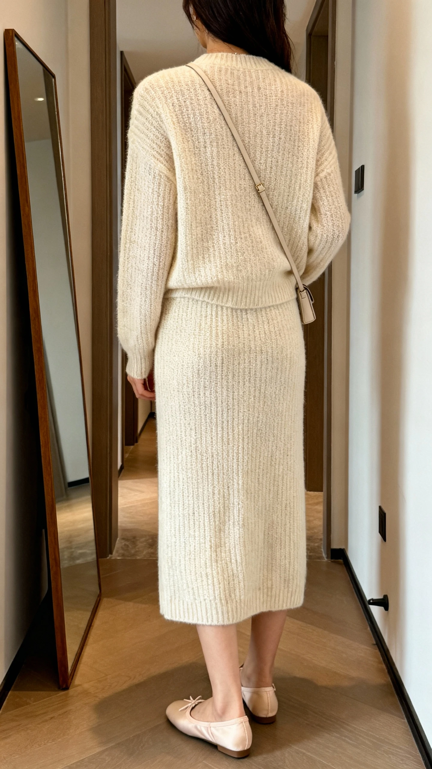 iPhone back view of a woman in a matching knit set (fine-knit sweater and coordinating knit midi skirt) with ballet flats and a minimal crossbody; face not visible, hallway full-length mirror, warm indoor lighting, iPhone photo quality.
