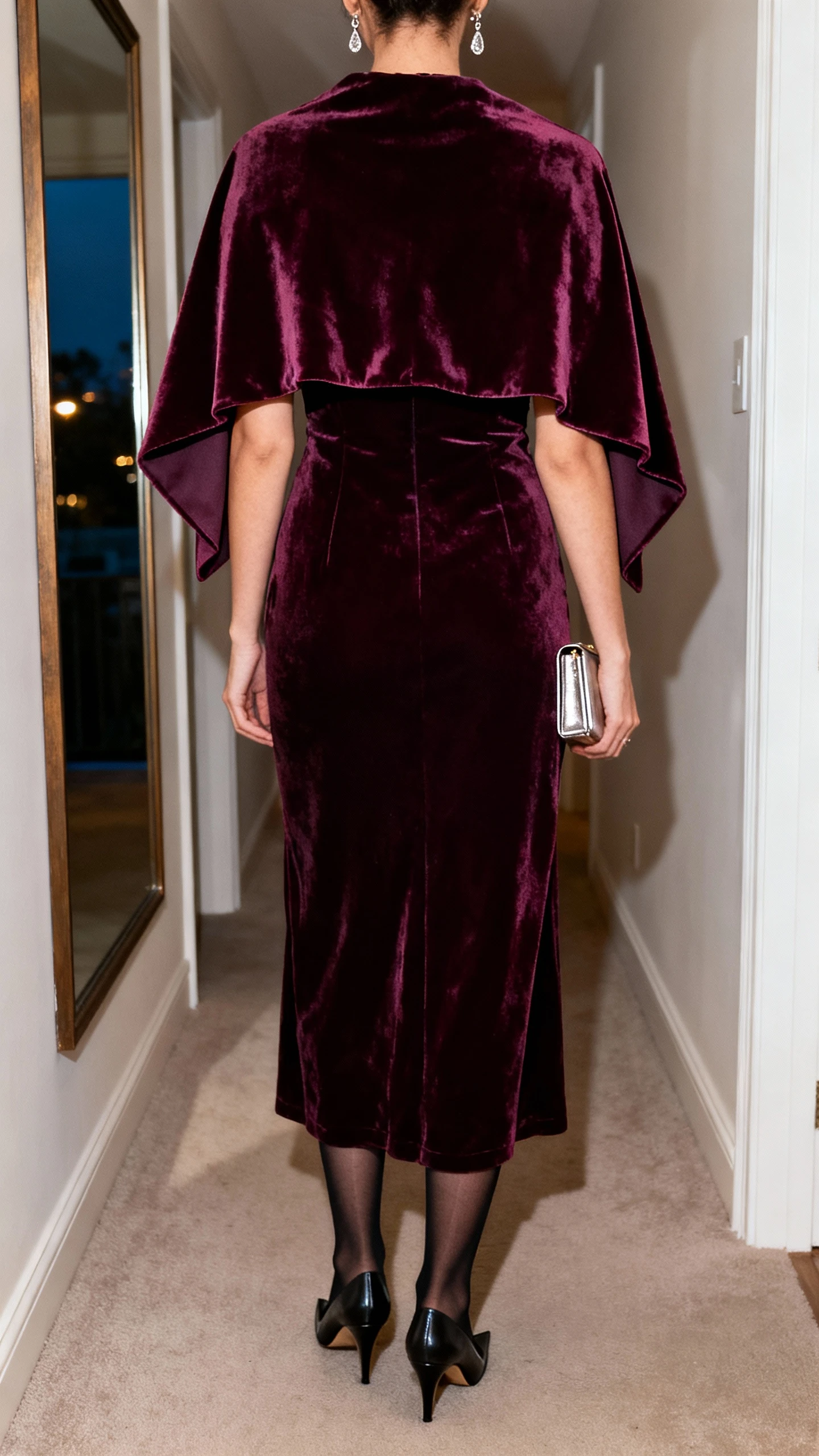 iPhone back view of a woman wearing a deep jewel-tone velvet maxi dress with a dramatic statement capelet, sheer black tights, pointed toe pumps, slim metallic clutch, and delicate drop earrings, face not visible, hallway mirror, indoor evening light, iPhone photo quality.