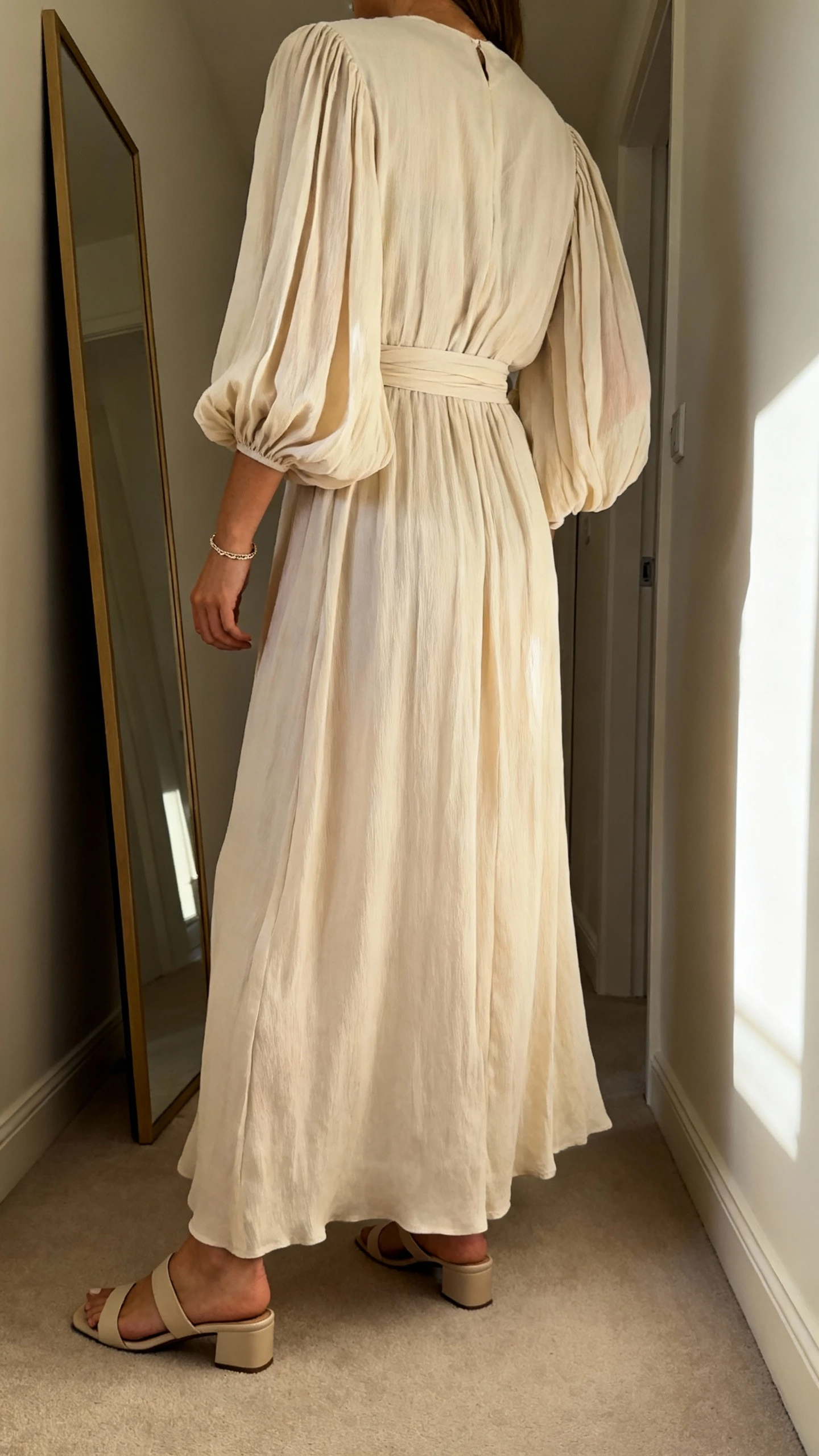 iPhone back view of a woman wearing a flowing maxi dress with dramatic long sleeves and a cinched waist, styled with low heeled sandals and a delicate bracelet, face not visible, hallway full-length mirror, morning light, casual iPhone photo.