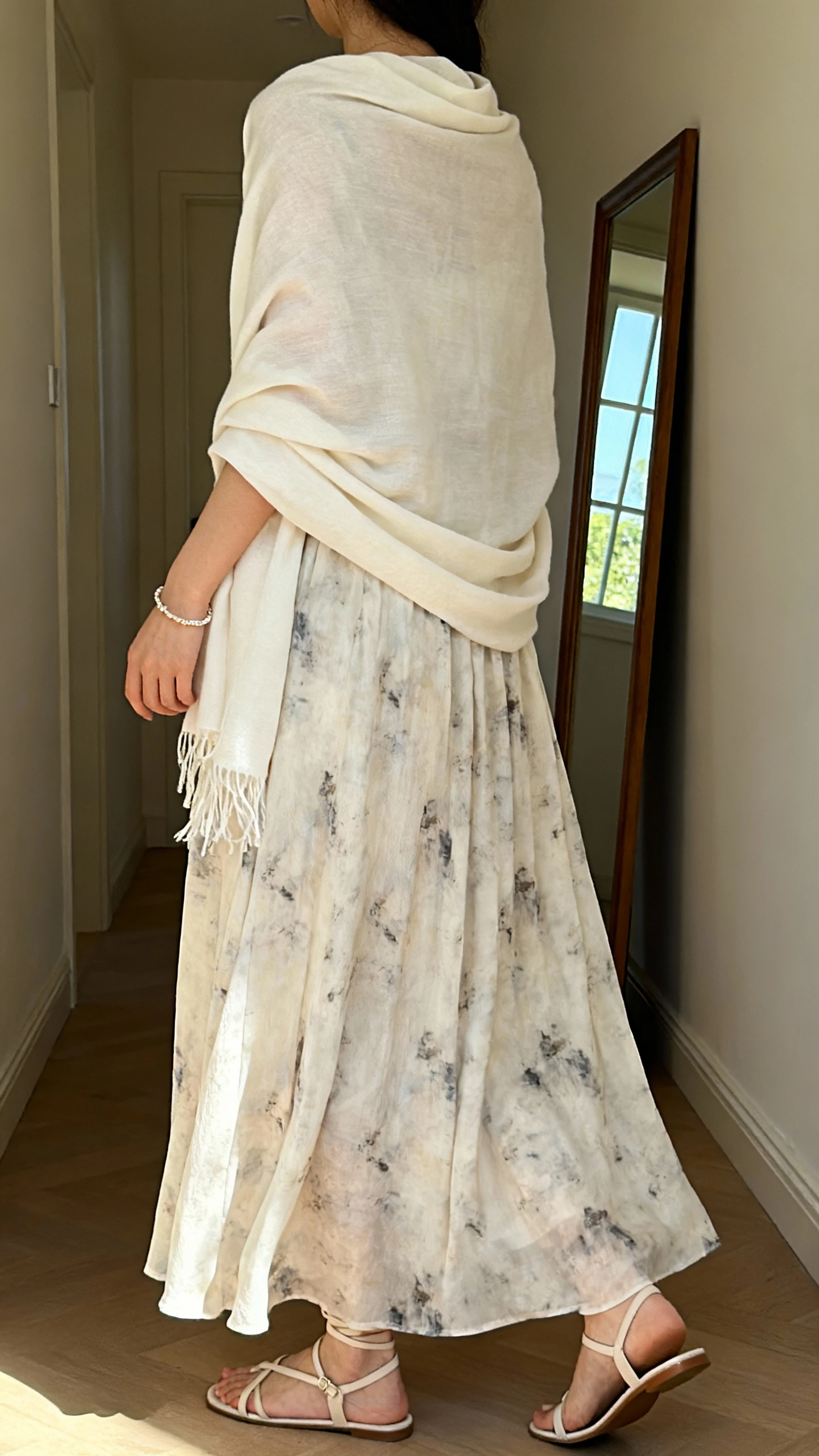 iPhone back view of a woman wearing a flowing maxi dress with subtle prints, light shawl draped over shoulders, delicate bracelet, and low strappy sandals, face not visible, hallway mirror, gentle afternoon window light, iPhone photo quality.