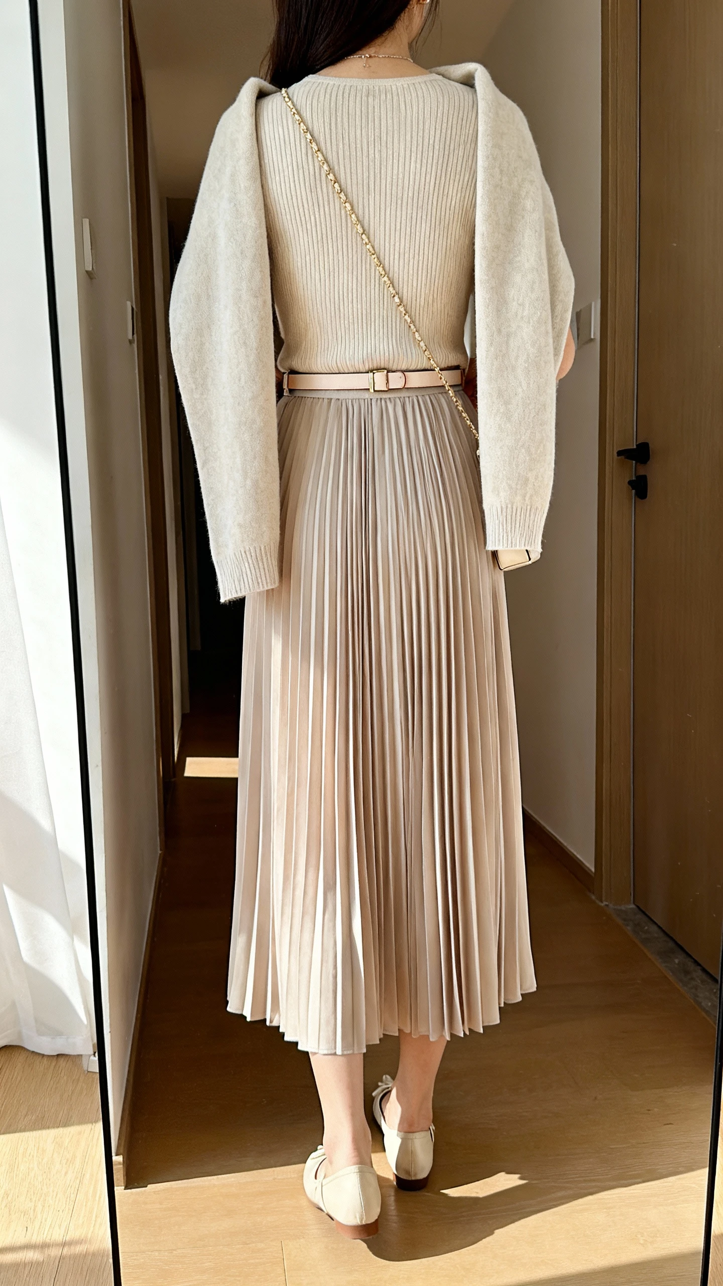 iPhone back view of a woman wearing a pleated midi skirt and fine-gauge knit top combo, soft cardigan draped over shoulders, dainty belt, ballet flats, and a small crossbody; face not visible, hallway mirror, indoor daylight, casual iPhone photo.