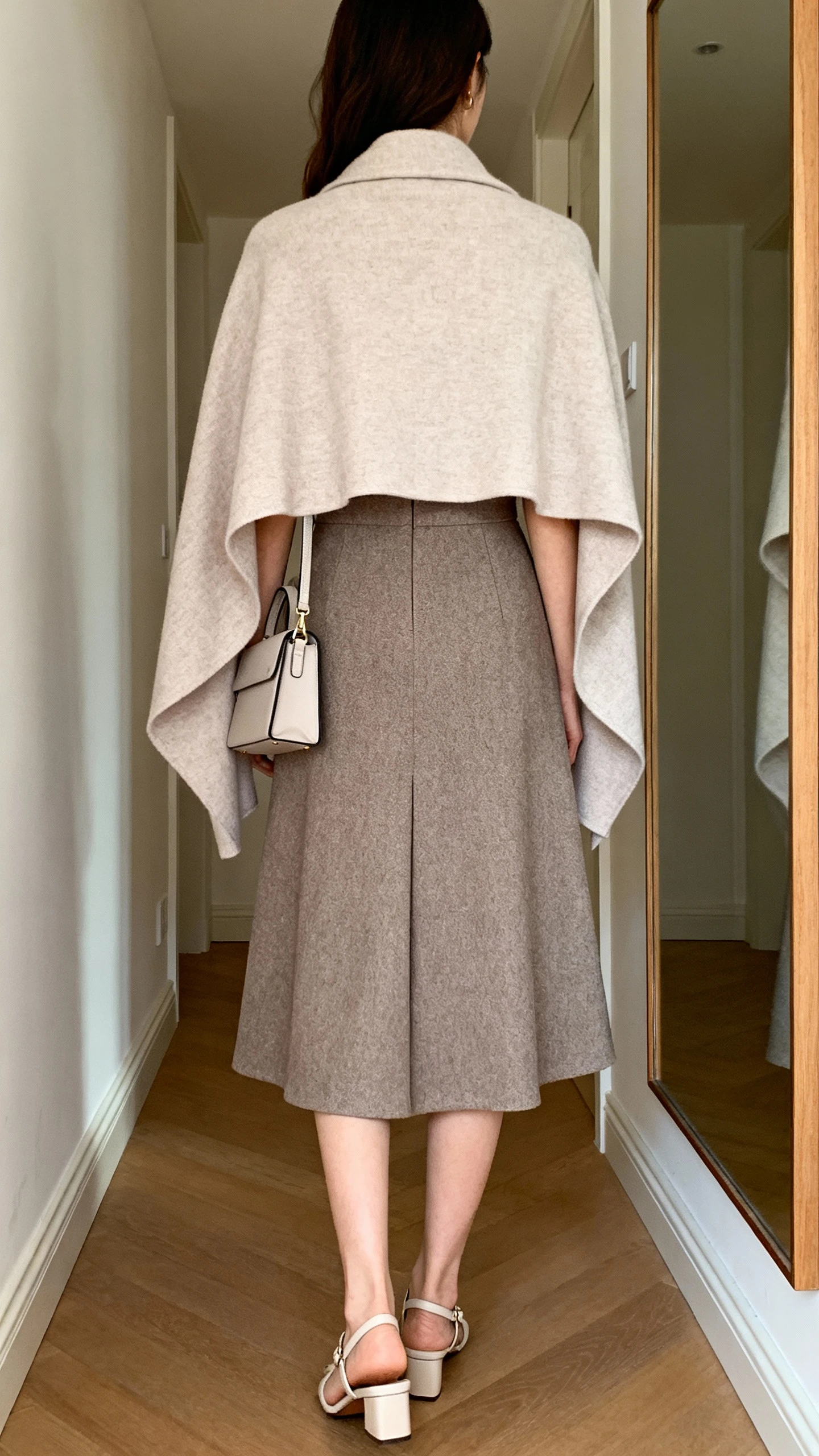 iPhone back view of a woman wearing a tailored A-line dress paired with a gentle cape or longline cardigan, simple closed-toe heels and a small structured bag, face not visible, hallway mirror, soft indoor lighting, iPhone photo quality