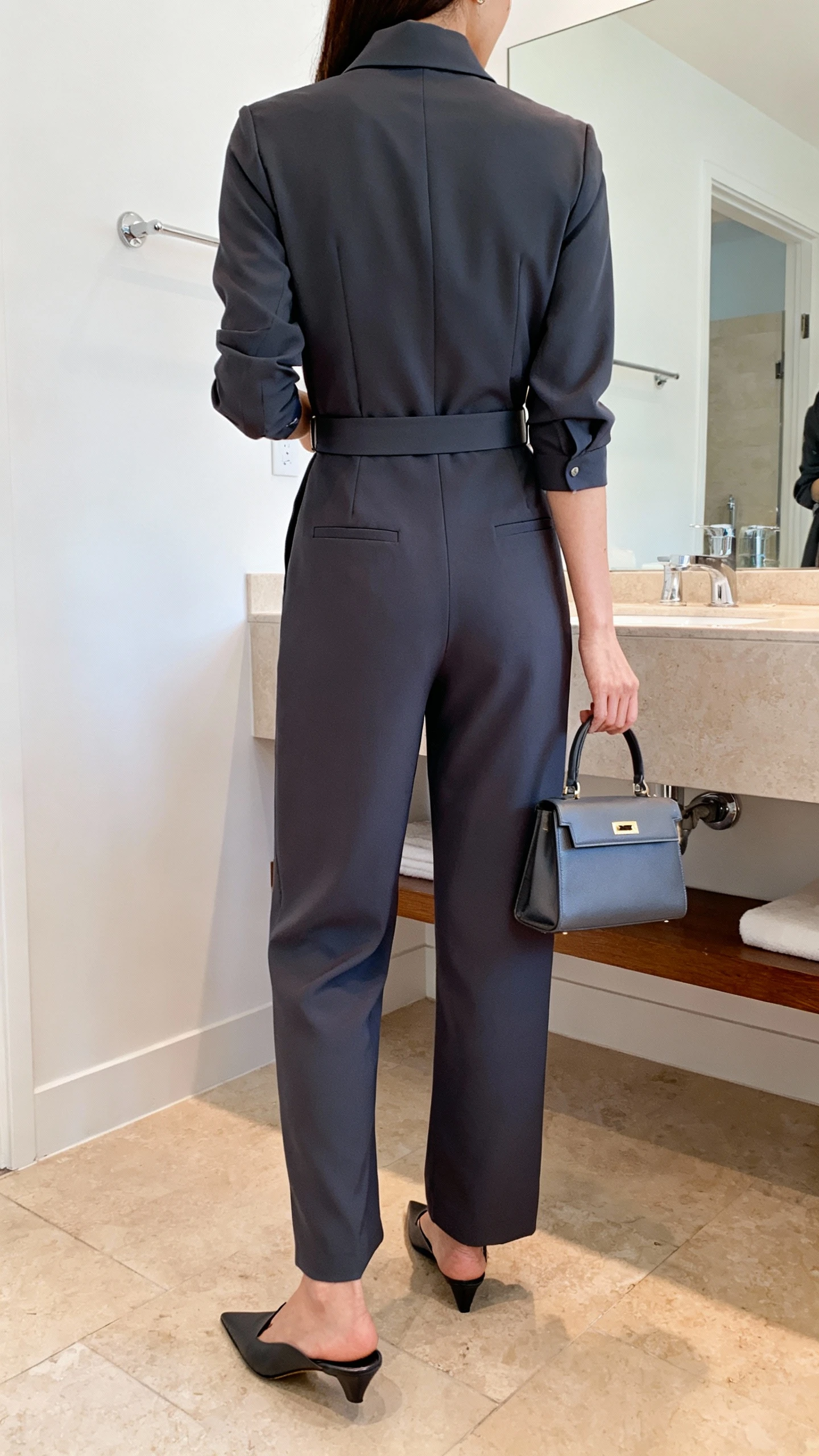 iPhone back view of a woman wearing a tailored jumpsuit with a poised high collar, slim waist belt, pointed flats, small structured top-handle bag, face not visible, bathroom mirror, bright indoor lighting, iPhone photo quality.