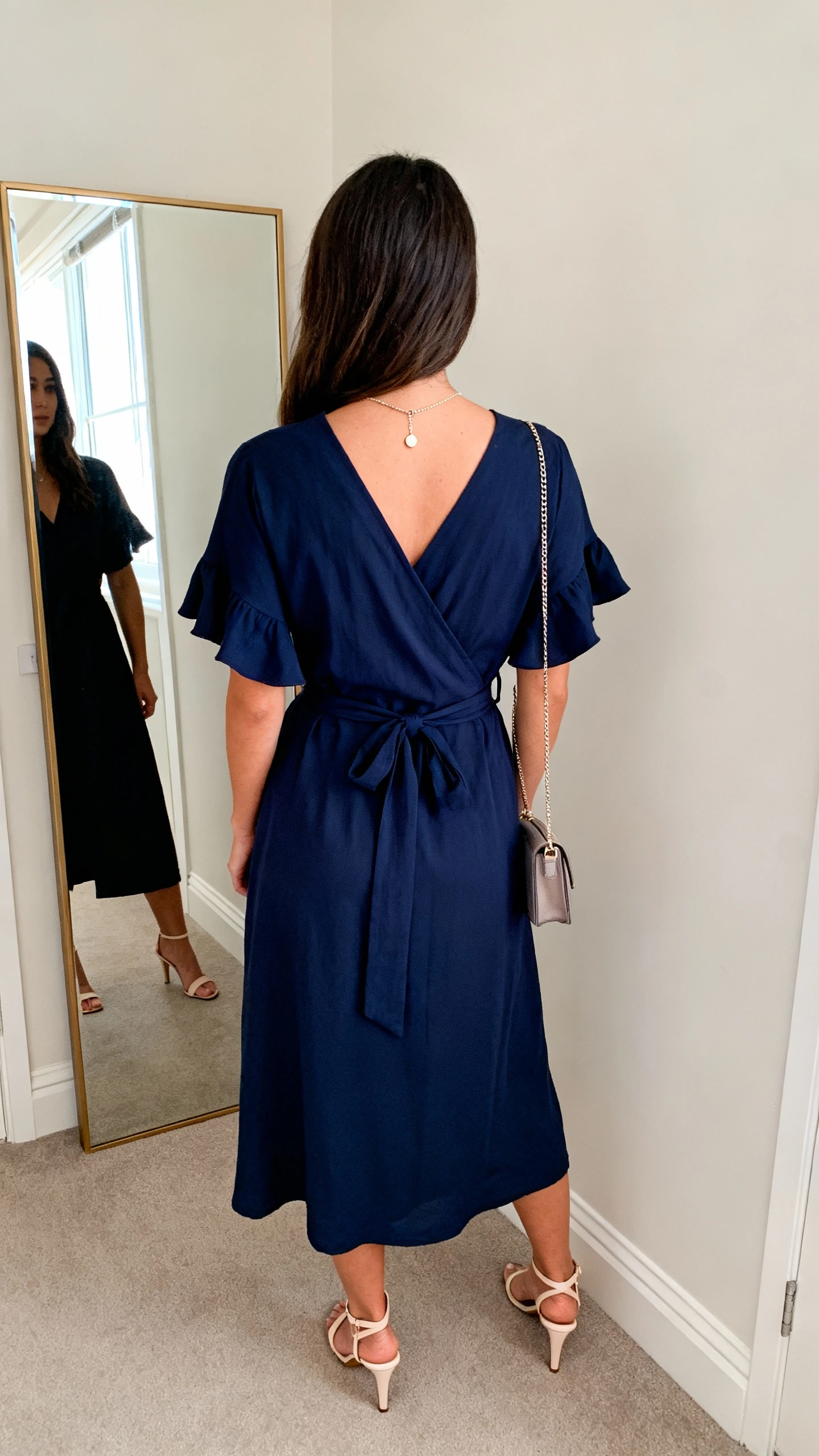 iPhone back view of a woman wearing a wrap dress in deep navy with a tie waist and flutter sleeves, dainty pendant necklace, small crossbody bag, and kitten-heel sandals, face not visible, hallway full-length mirror, natural daylight from a window, iPhone photo quality.