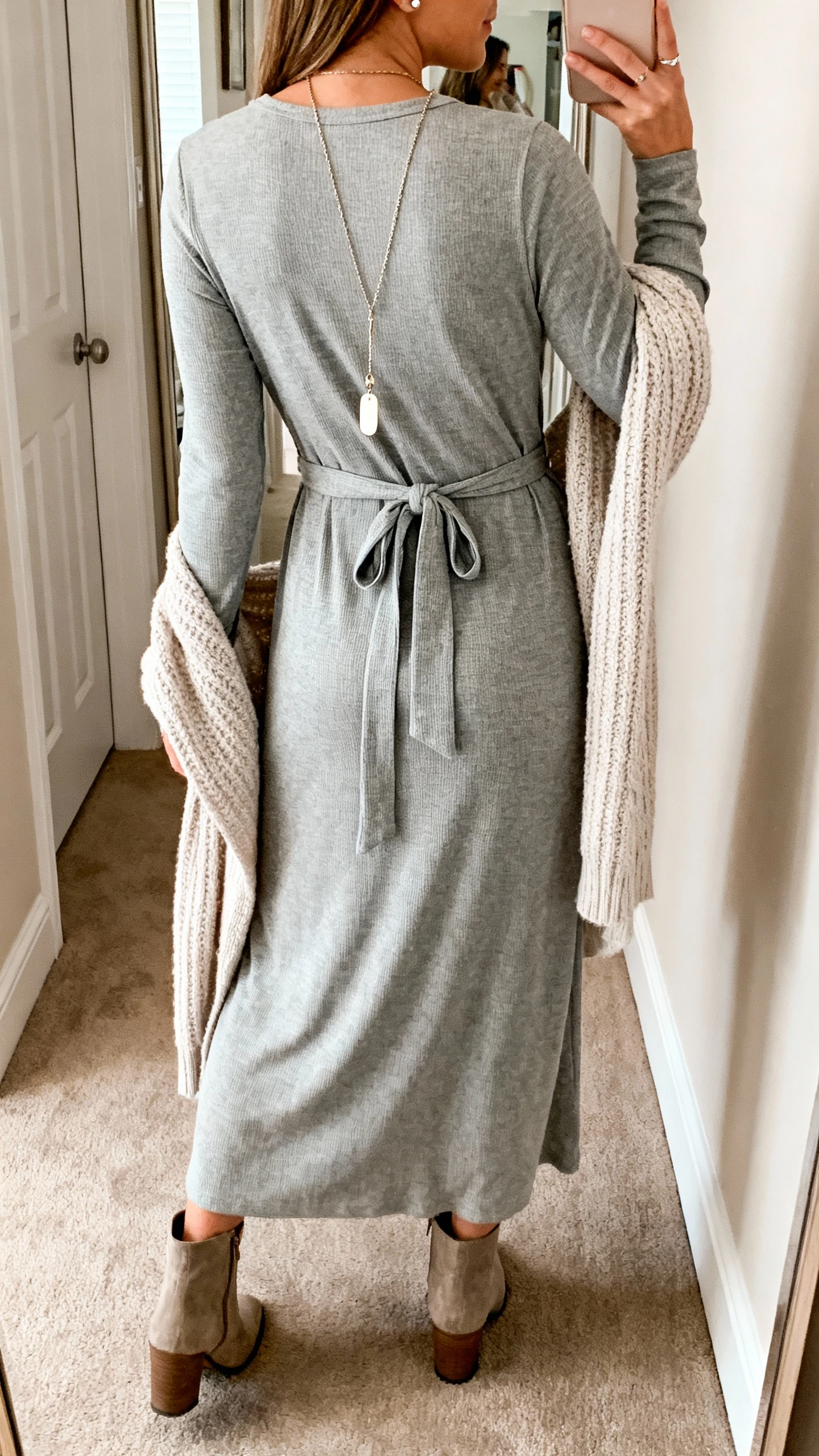 iPhone back-view photo of a woman wearing a relaxed-fit jersey maxi dress with long sleeves, a soft self-tie belt loosely at the natural waist, low-heel ankle boots, a long pendant necklace with simple studs, and a knitted duster draped over her shoulders; face not visible, hallway mirror, natural daylight, iPhone photo quality.