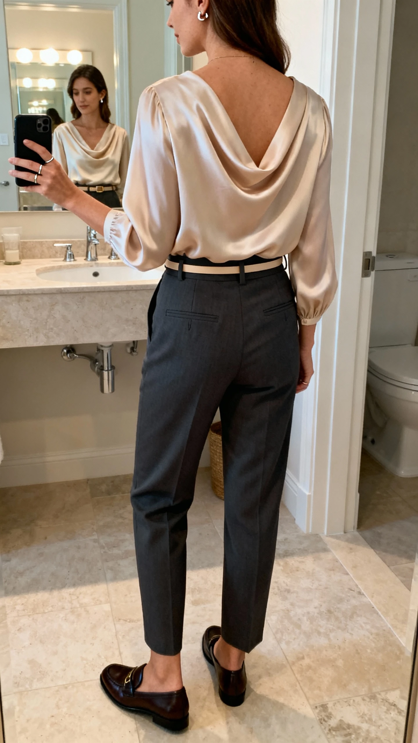iPhone back-view photo of a woman wearing high-waisted tailored trousers with a drape-front silk blouse, classic loafers, a thin belt, subtle stud earrings, and a slender ring, face not visible, bathroom mirror with indoor lighting, iPhone photo quality.