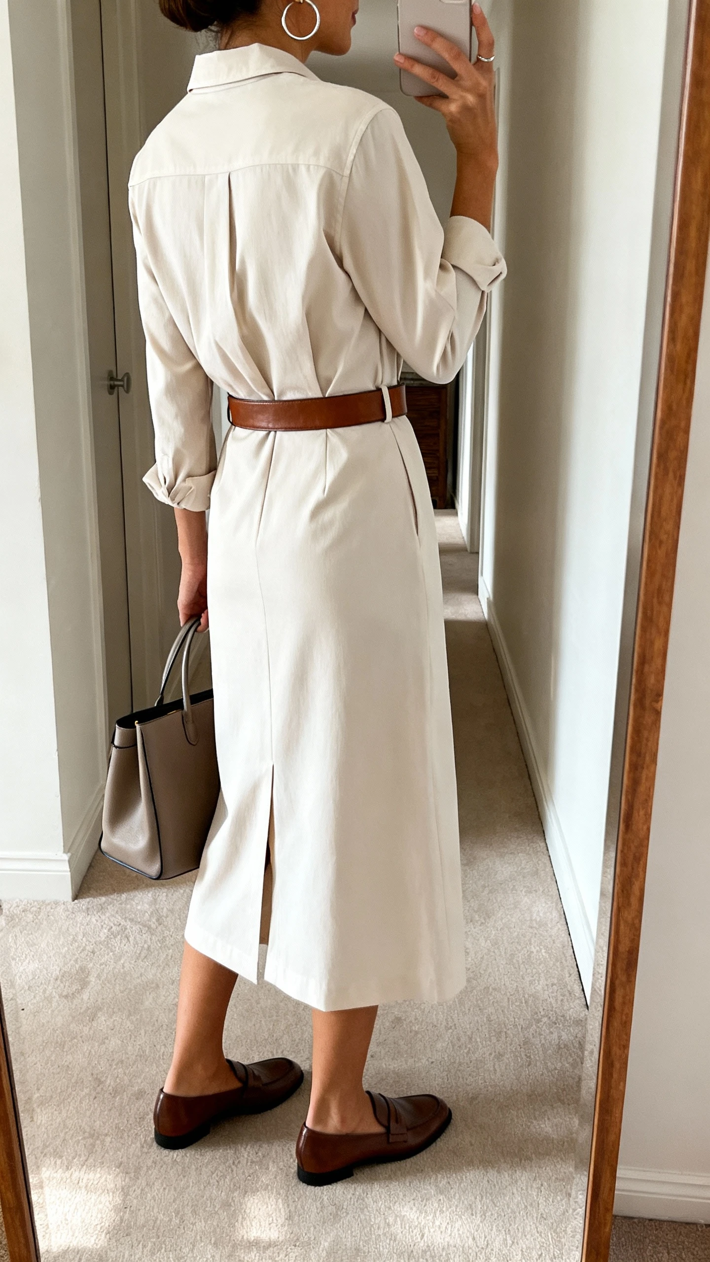 iPhone back-view selfie of a woman wearing a tailored midi shirtdress with a gentle fit, leather waist belt, loafers, a structured tote, and simple hoop earrings, face not visible, hallway mirror, indoor daylight, iPhone photo quality.
