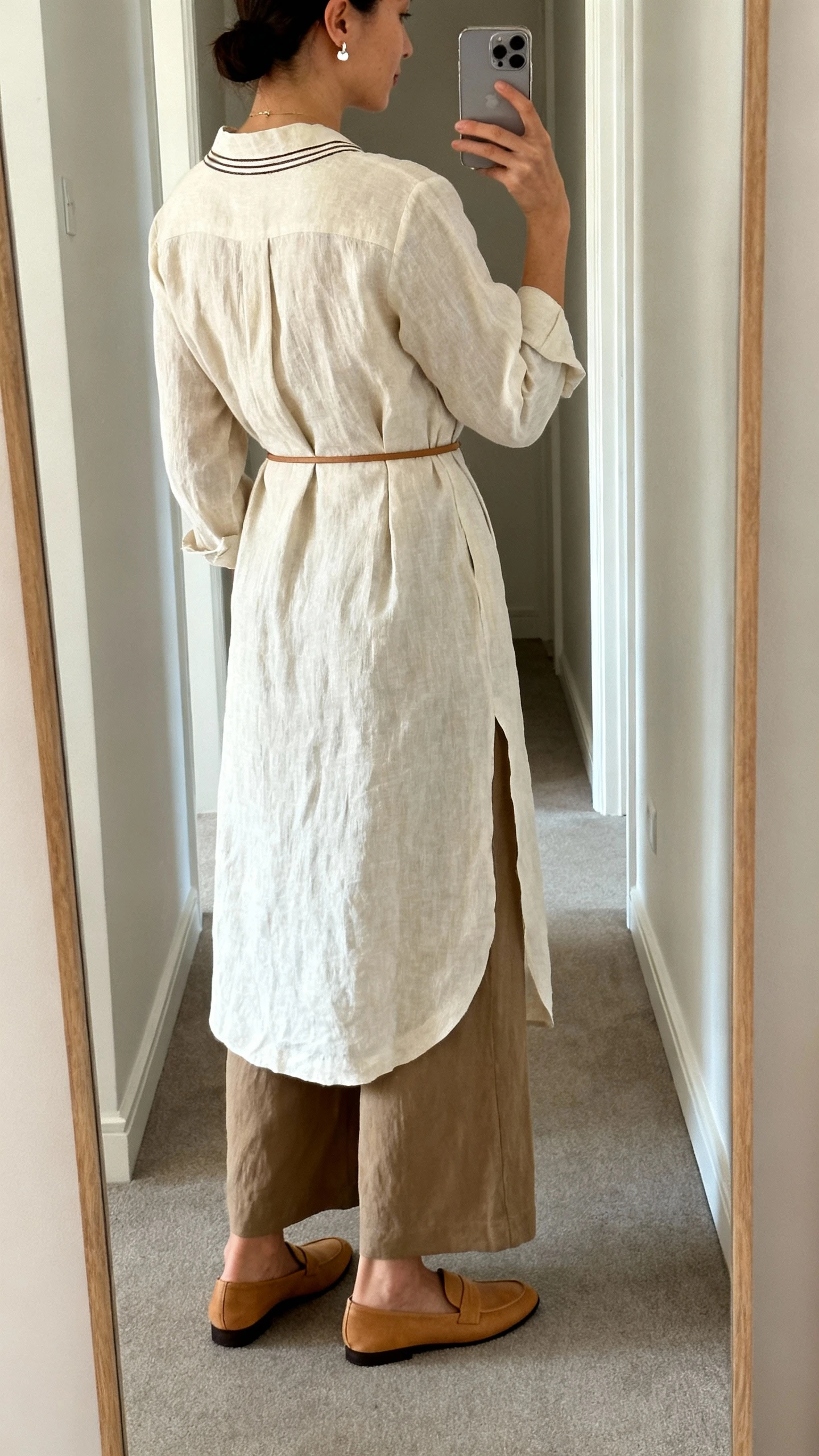 iPhone back-view selfie of a woman wearing high-rise wide-leg trousers in drapey linen-blend, a longline tunic shirt with a banded neckline, tan loafers, a thin waist-cinching belt over the tunic, and minimal stud earrings with a subtle pendant, face not visible, hallway mirror, natural daylight, iPhone photo quality.