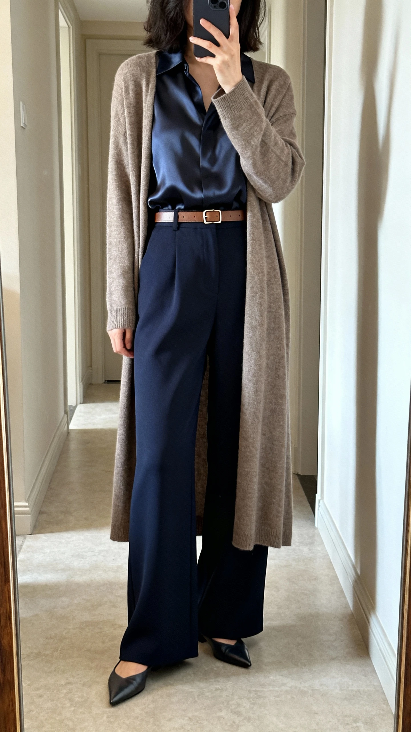 iPhone mirror selfie back view of a woman wearing high-waisted wide-leg trousers in deep navy, a satin-effect blouse, a complementary neutral longline cardigan, pointed-toe flats, and a minimalist leather belt with a small buckle, face not visible, full-length hallway mirror with natural daylight, iPhone photo quality.