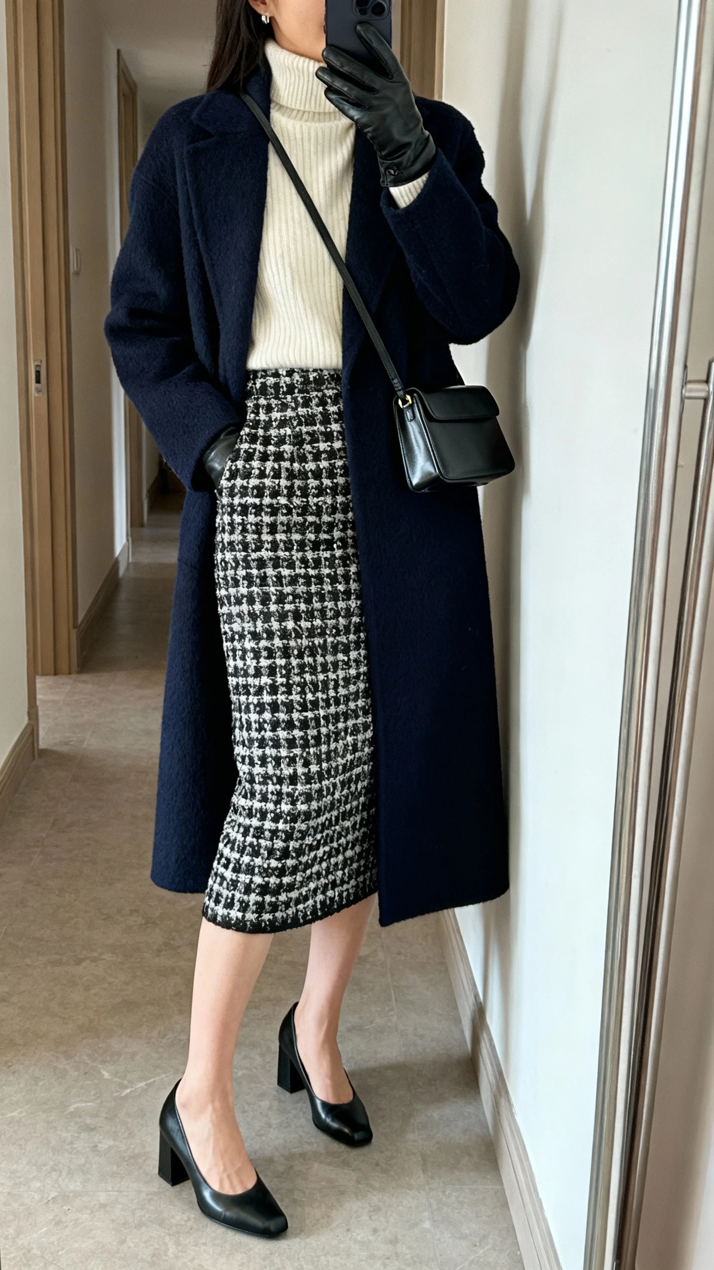 iPhone mirror selfie of a woman in a black-and-white tweed midi pencil skirt, cream fine-knit turtleneck, closed-toe black block-heel pumps, longline navy wool coat, and leather gloves with a slim leather shoulder bag, face not visible, side angle in a hallway full-length mirror, indoor natural light, iPhone photo quality.