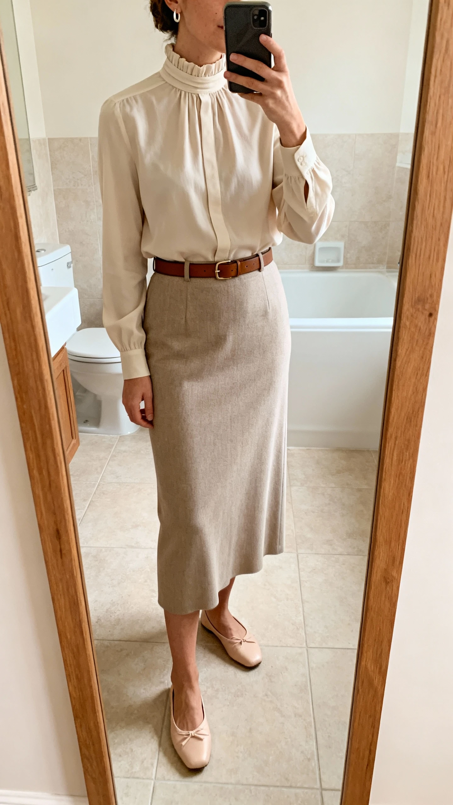 iPhone mirror selfie of a woman in a column skirt and a high-neck blouse tucked in, slim leather belt, ballet flats, minimal earrings, face not visible, cropped at neck, bathroom mirror, soft indoor lighting, iPhone photo quality.