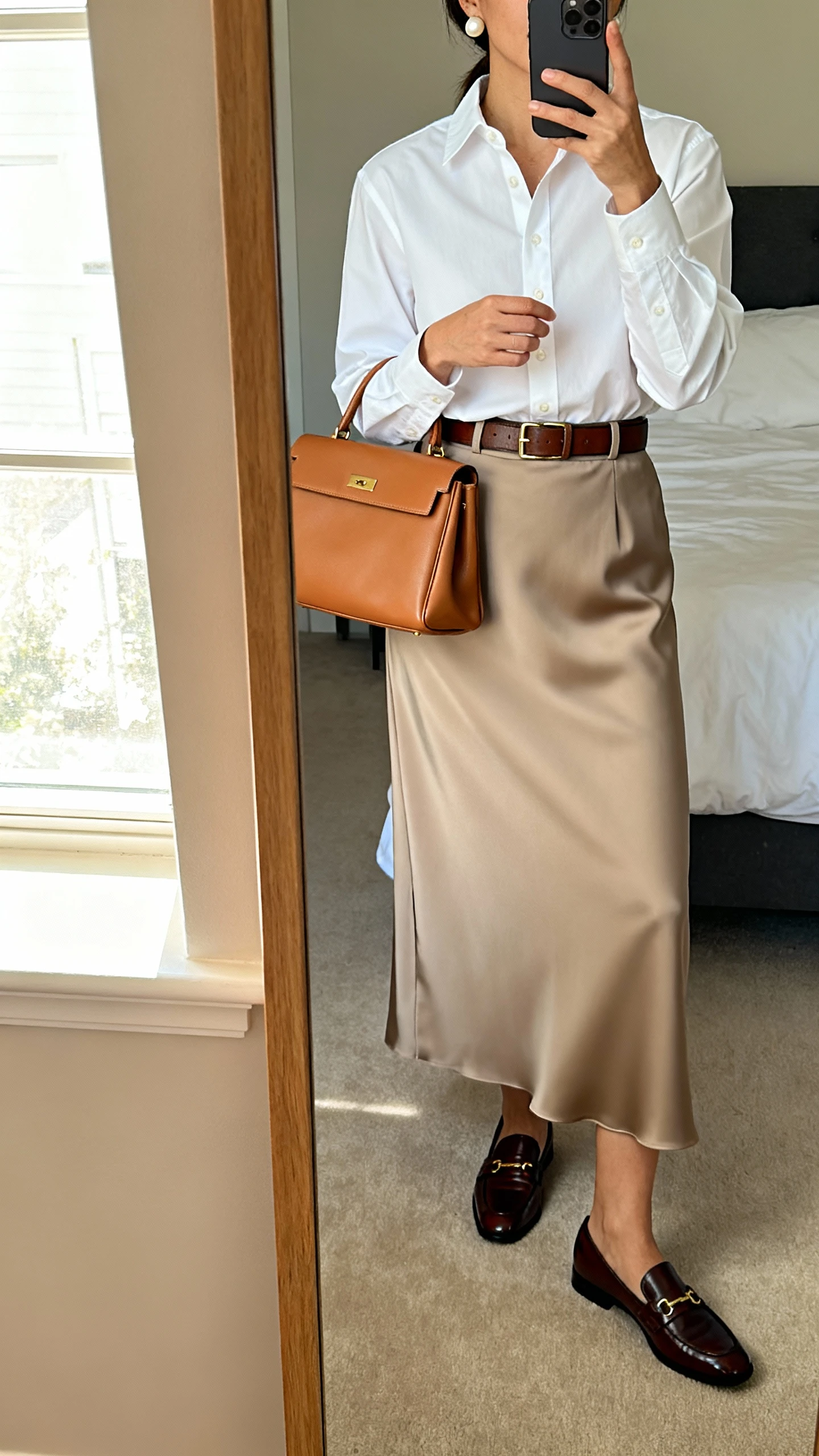 iPhone mirror selfie of a woman in a crisp white button-down tucked into a high-waisted crepe maxi skirt, polished loafers, an optional leather belt, a structured tan handbag, and pearl studs, face not visible, cropped at shoulders, bedroom mirror with window light, iPhone photo quality.