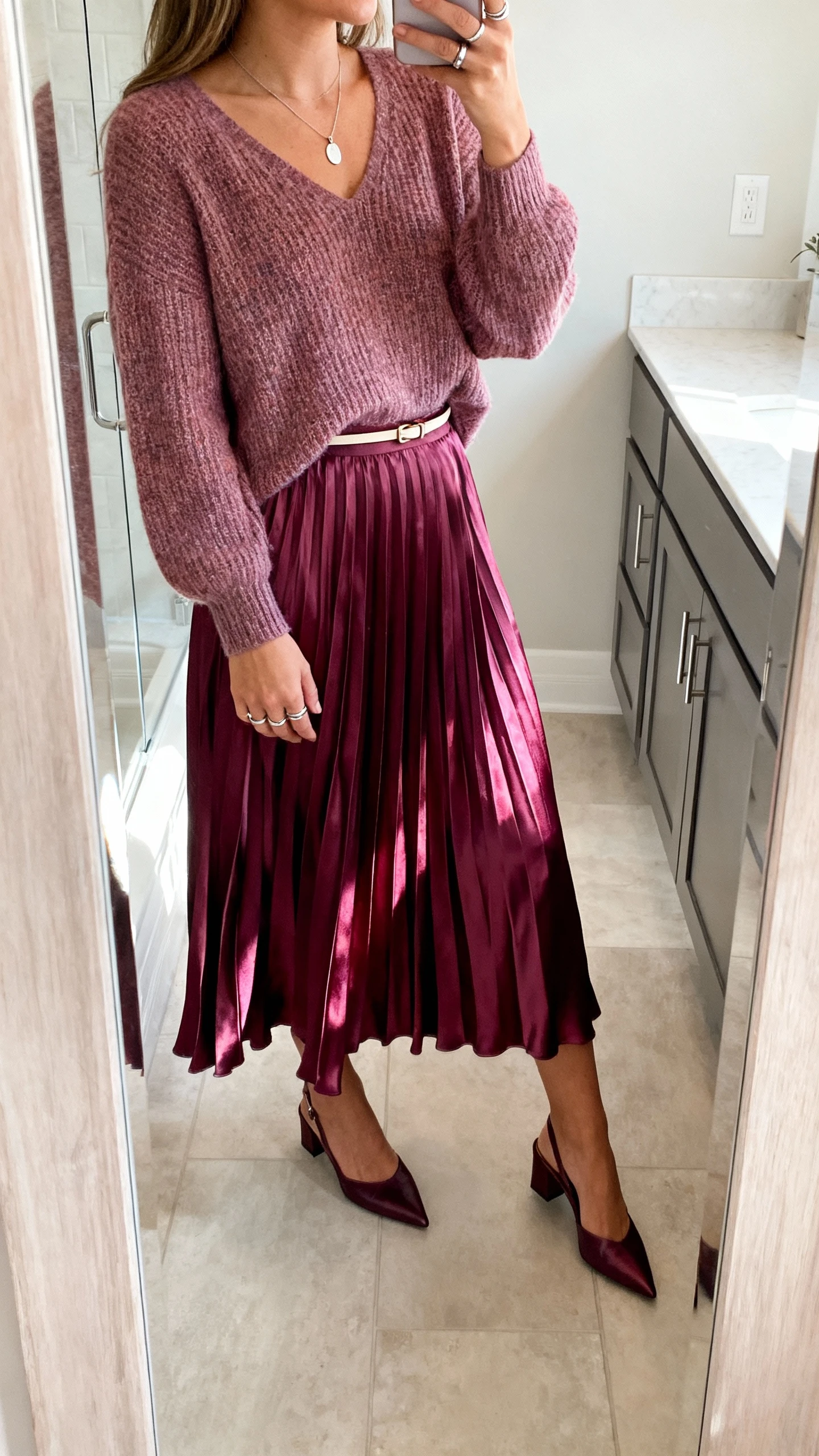 iPhone mirror selfie of a woman in a flowy pleated satin midi skirt (jewel tone) with a lightweight knit sweater with a modest neckline, pointed slingback flats, a thin waist belt, a delicate pendant necklace and stacked rings, face not visible, side angle in a bathroom mirror, soft morning light, iPhone photo quality.