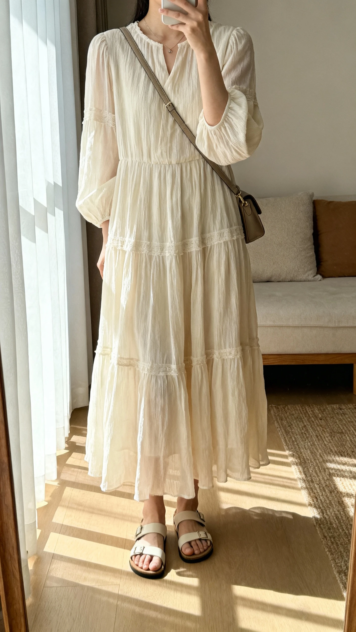 iPhone mirror selfie of a woman in a flowy tiered maxi dress with sleeves and full coverage, light airy fabric, paired with simple sandals and a crossbody bag, face not visible, cropped at neck, living room mirror, gentle afternoon window light, iPhone photo quality.