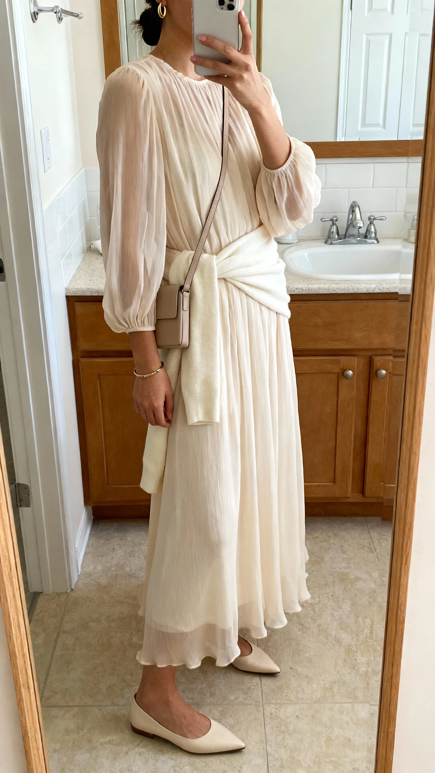 iPhone mirror selfie of a woman in a long-sleeve chiffon maxi dress with a gentle A-line, a soft cream wrap, pointed-toe flats, a slim neutral crossbody, small gold hoops, and a simple bracelet; face not visible, side angle, bathroom mirror, morning indoor lighting, iPhone photo quality.
