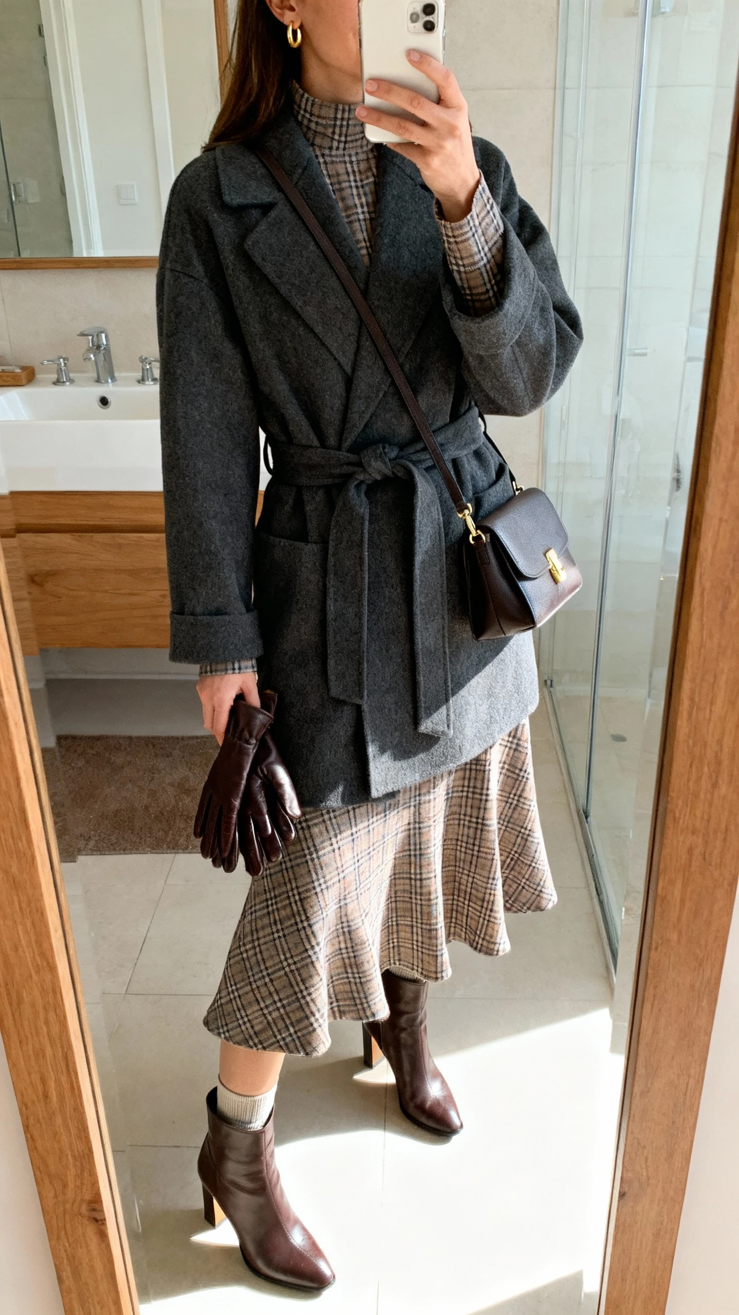 iPhone mirror selfie of a woman in a long-sleeve plaid midi dress with a gently flared skirt, belted wrap coat in charcoal, heeled leather ankle boots, thermal slip underneath for warmth, leather gloves tucked in hand, structured crossbody bag, and small gold hoop earrings; face not visible, bathroom mirror, indoor morning light, iPhone photo quality.