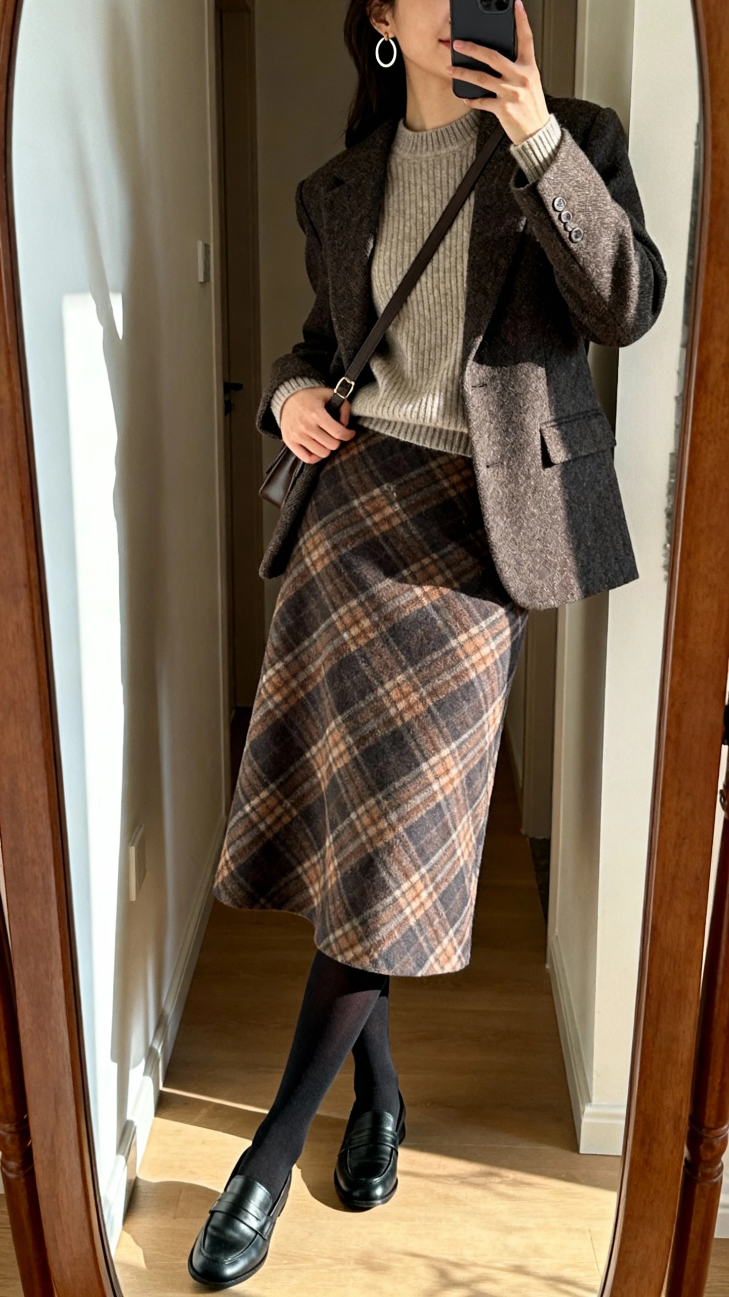 iPhone mirror selfie of a woman in a midi plaid A-line skirt, fitted crewneck sweater, tailored sweater-blazer layered on top, black opaque tights, loafers, simple hoop earrings, crossbody bag, face not visible, side angle in hallway full-length mirror, indoor natural light, iPhone photo quality.