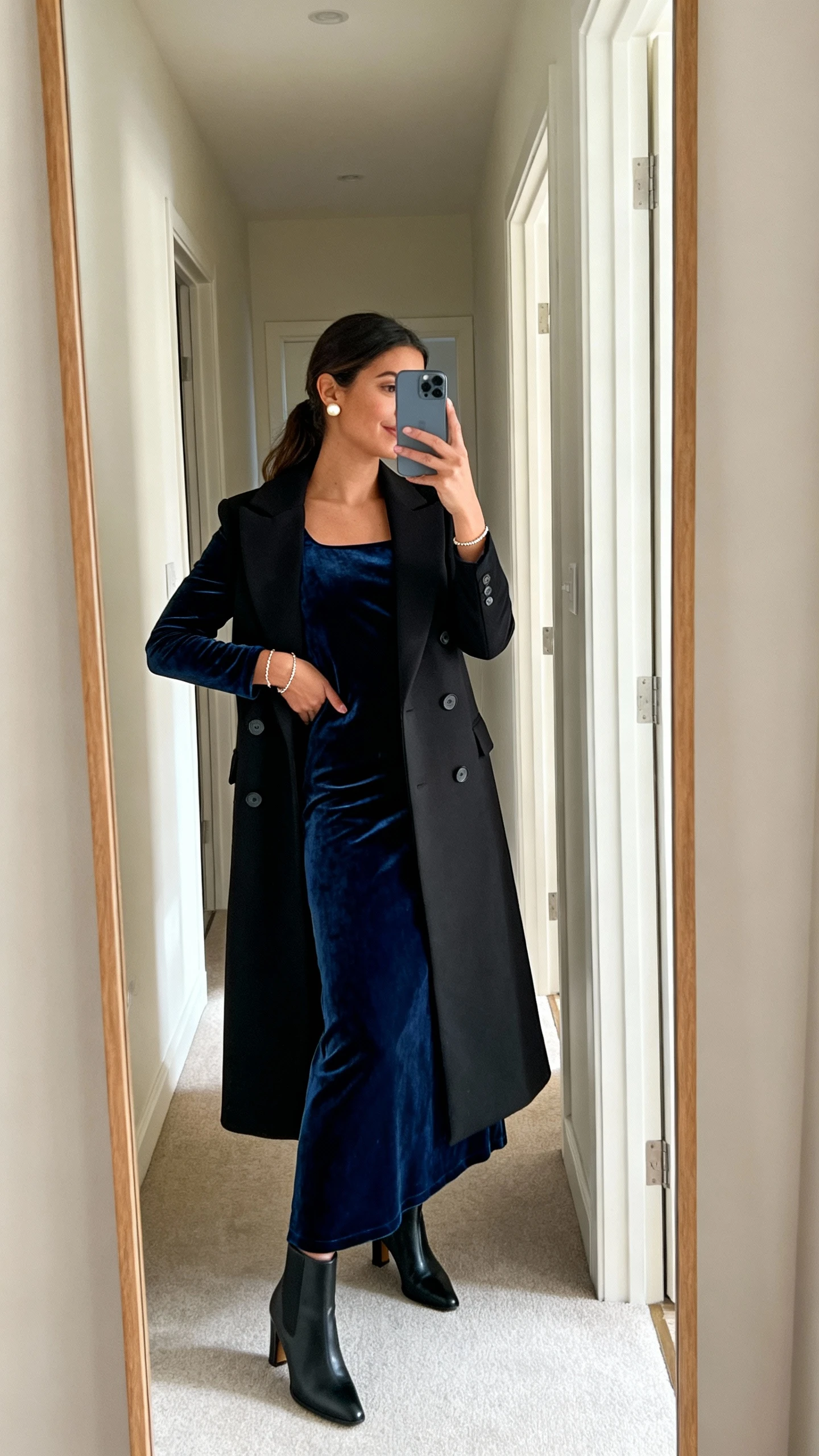 iPhone mirror selfie of a woman in a midnight blue velvet long-sleeve maxi dress with a boat neckline, black double-breasted tailored long coat, black heeled ankle boots, pearl stud earrings and a delicate bracelet, face not visible, side angle in hallway mirror, soft indoor morning light, iPhone photo quality.