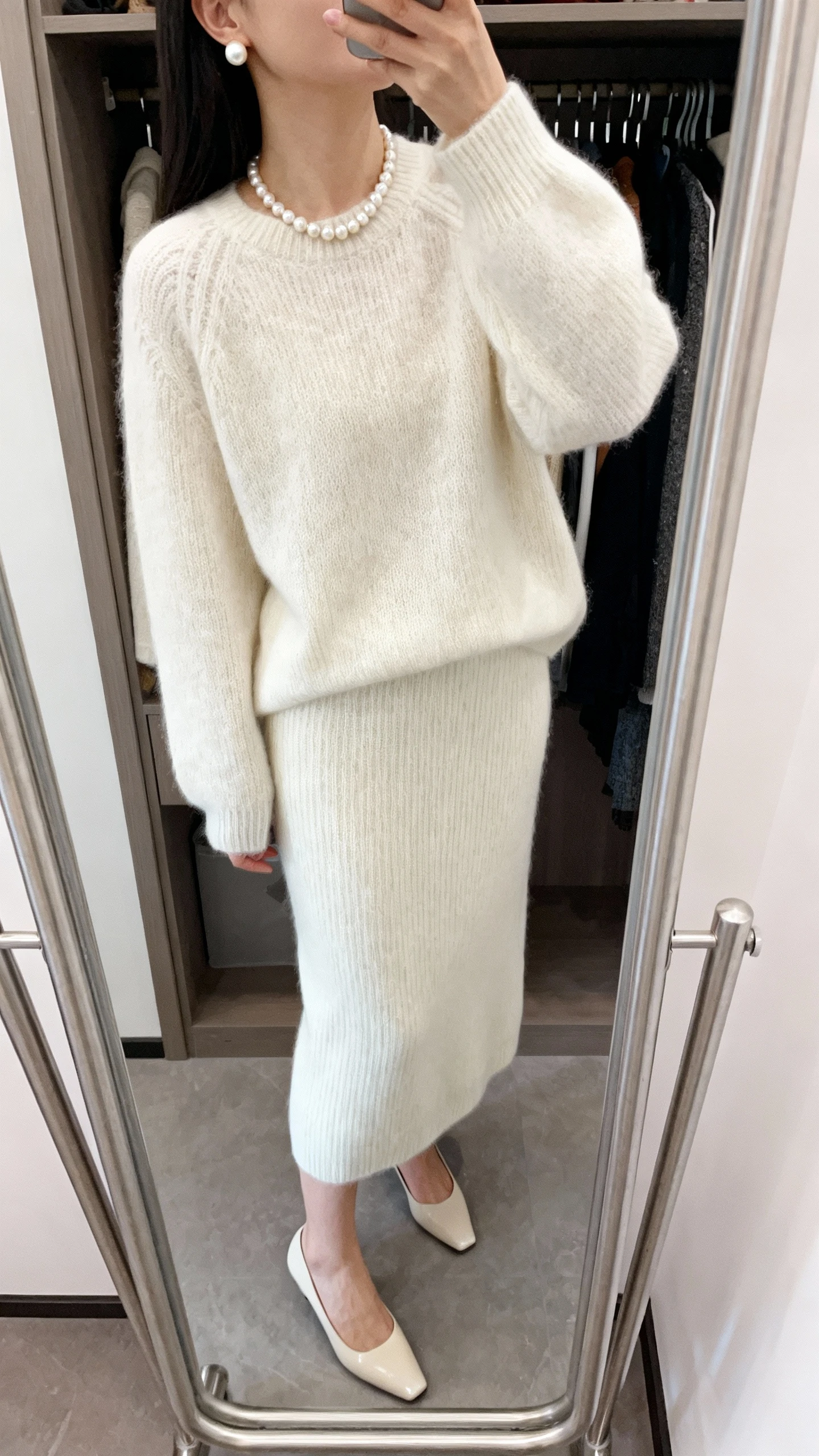 iPhone mirror selfie of a woman in a monochrome knit set (matching sweater and skirt) with a pearl necklace, pearl stud earrings, and sleek low heels, face not visible, cropped at neck, closet mirror, gentle indoor light, iPhone photo quality