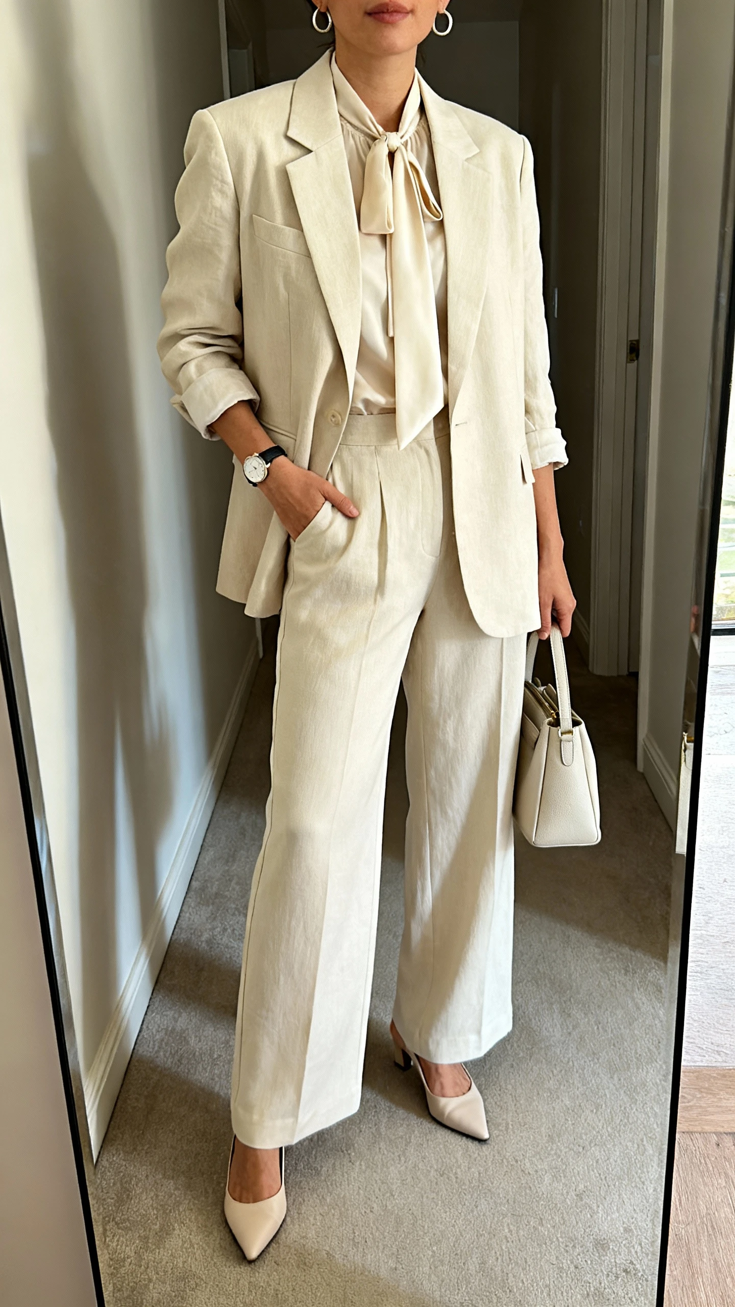 iPhone mirror selfie of a woman in a monochrome suit: single-breasted blazer with slightly relaxed fit, matching high-waisted wide-leg trousers, silk tie-neck blouse, low kitten heels, minimalist watch, medium huggie earrings, and a structured bag, face not visible, cropped at shoulders, hallway full-length mirror, natural daylight, iPhone photo quality.