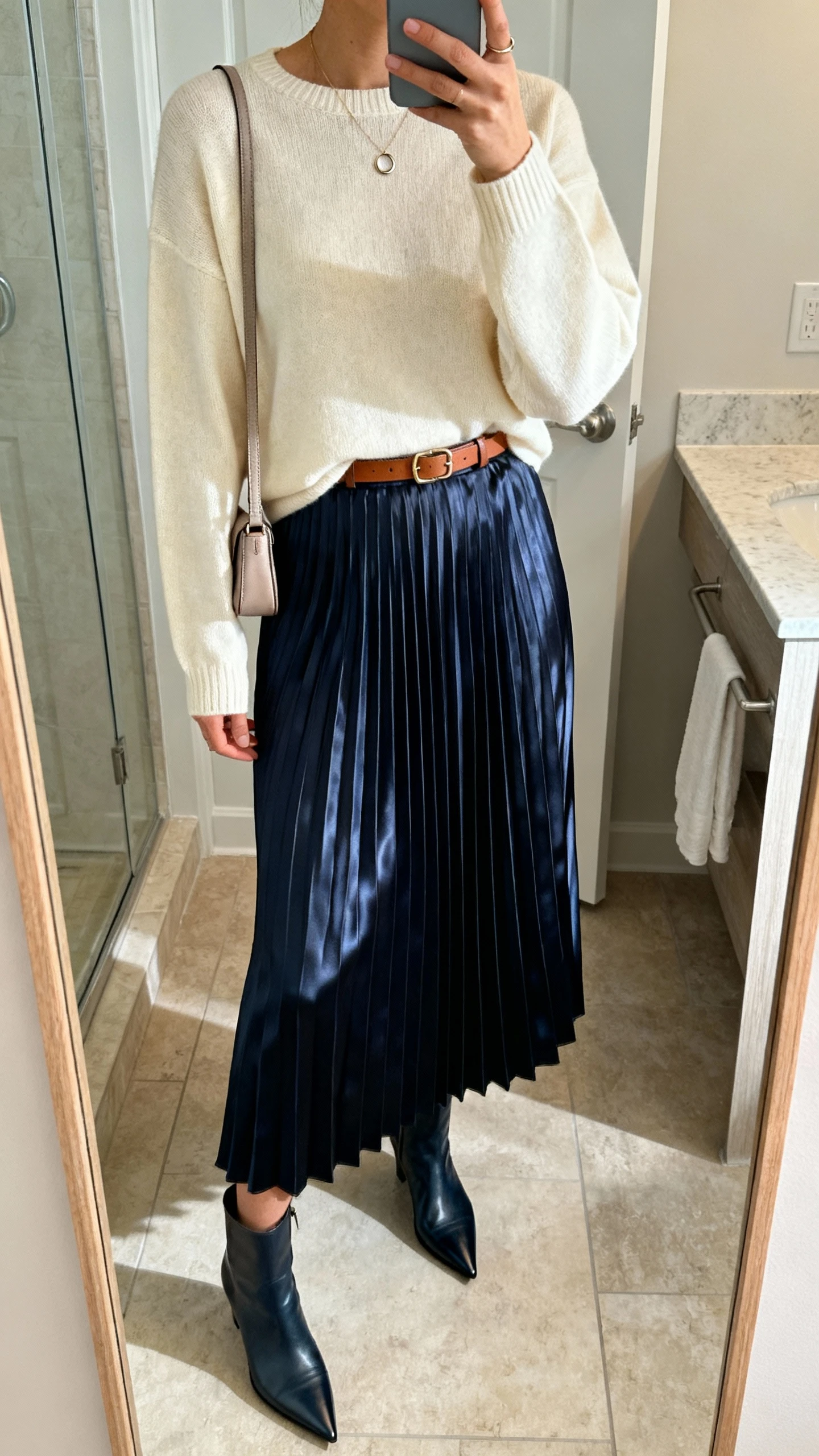 iPhone mirror selfie of a woman in a navy satin pleated midi skirt, cream lightweight crewneck sweater, pointed-toe ankle boots, a thin leather belt, a petite shoulder bag, and a minimal pendant necklace with small hoops; face not visible, bathroom mirror, indoor lighting, iPhone photo quality.