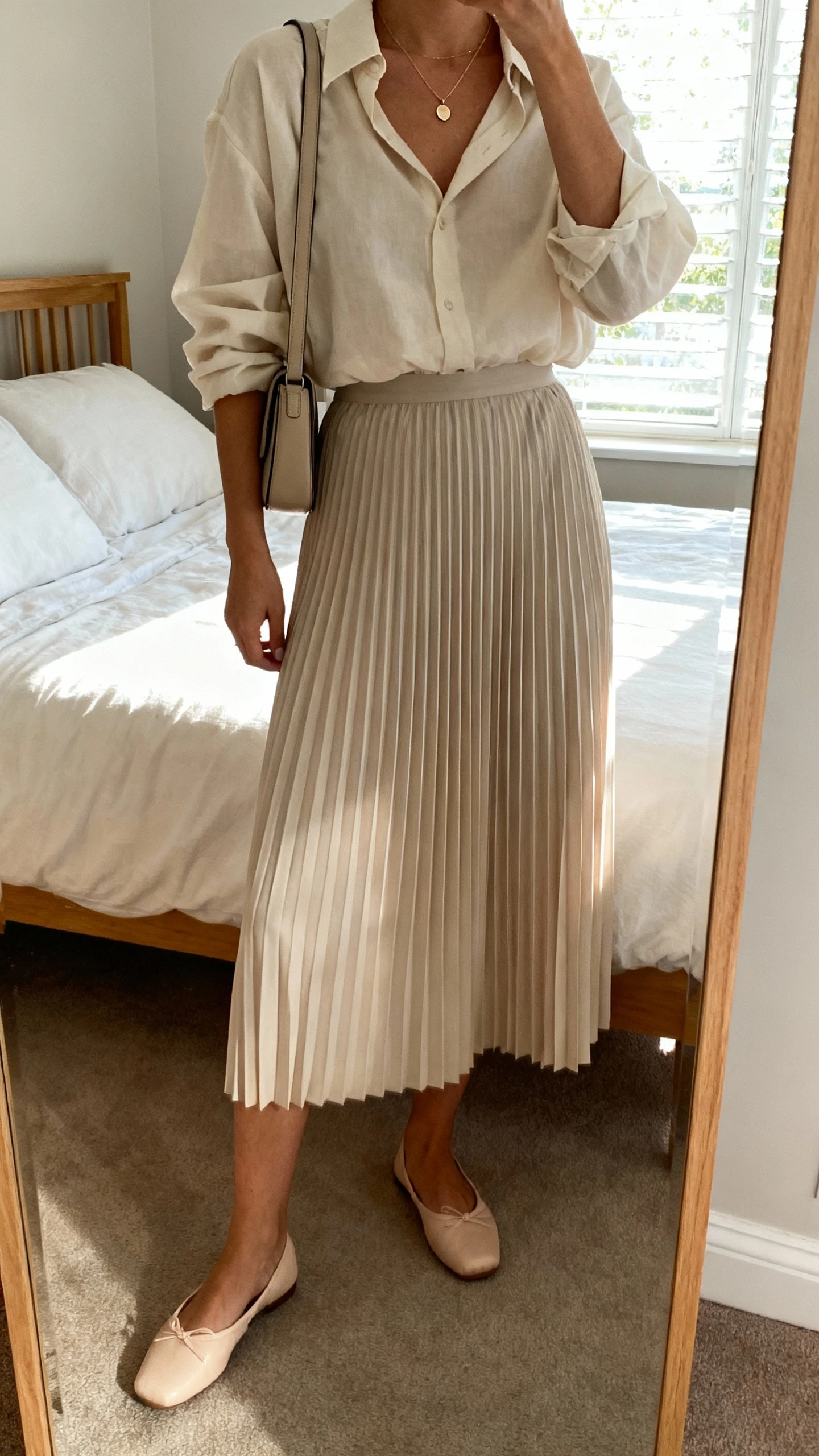 iPhone mirror selfie of a woman in a pleated midi skirt and relaxed blouse tucked loosely, ballet flats, a slim shoulder bag, and a dainty pendant necklace, face not visible, cropped at shoulders, bedroom mirror with natural window light, iPhone photo quality.