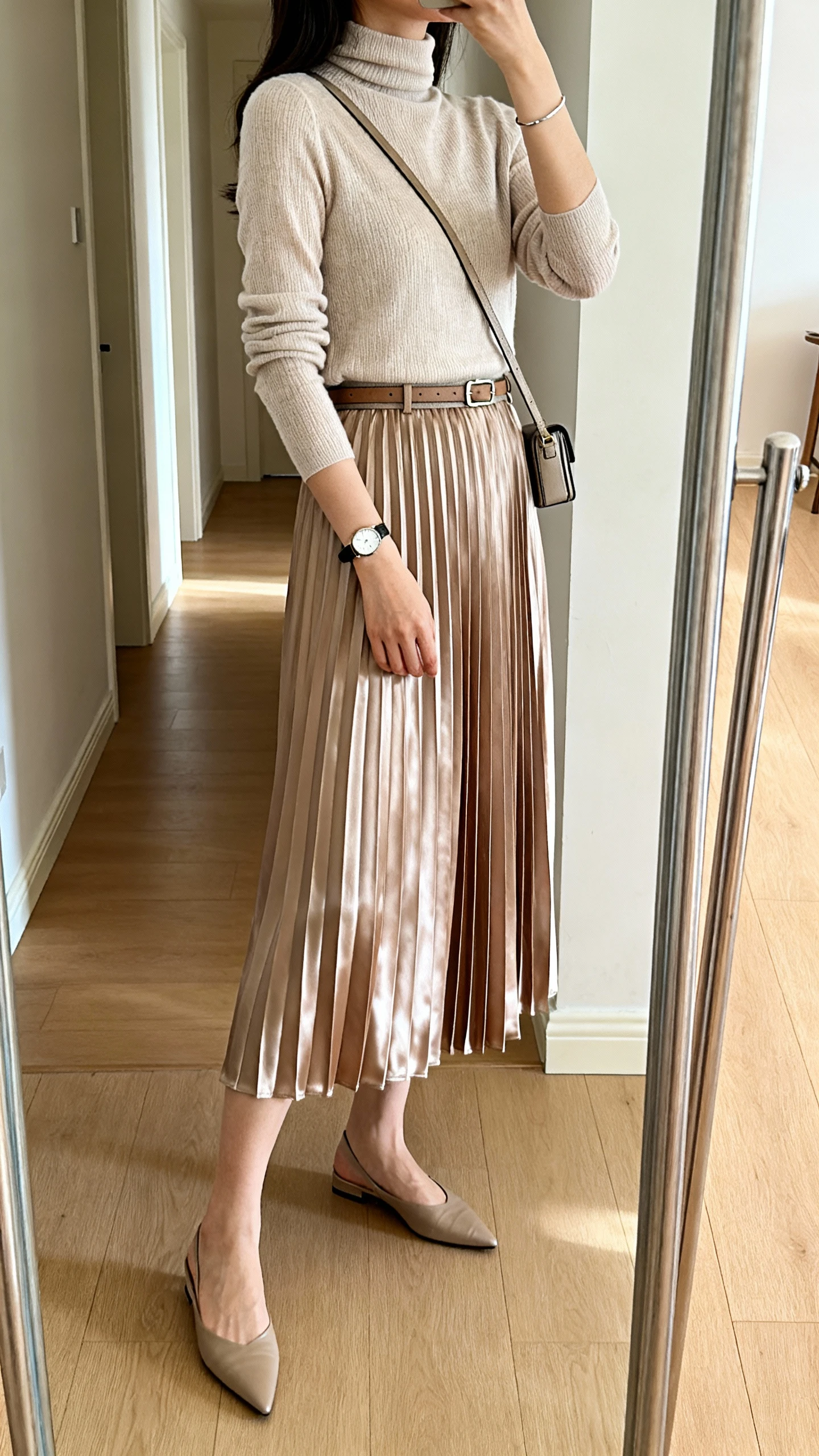 iPhone mirror selfie of a woman in a satin midi pleated skirt with a lightweight mock-neck knit top, pointed flats, a simple watch, slim bracelet, optional waistbelt, and a compact crossbody with a strap, face not visible, side angle in a full-length hallway mirror, soft morning light, iPhone photo quality.