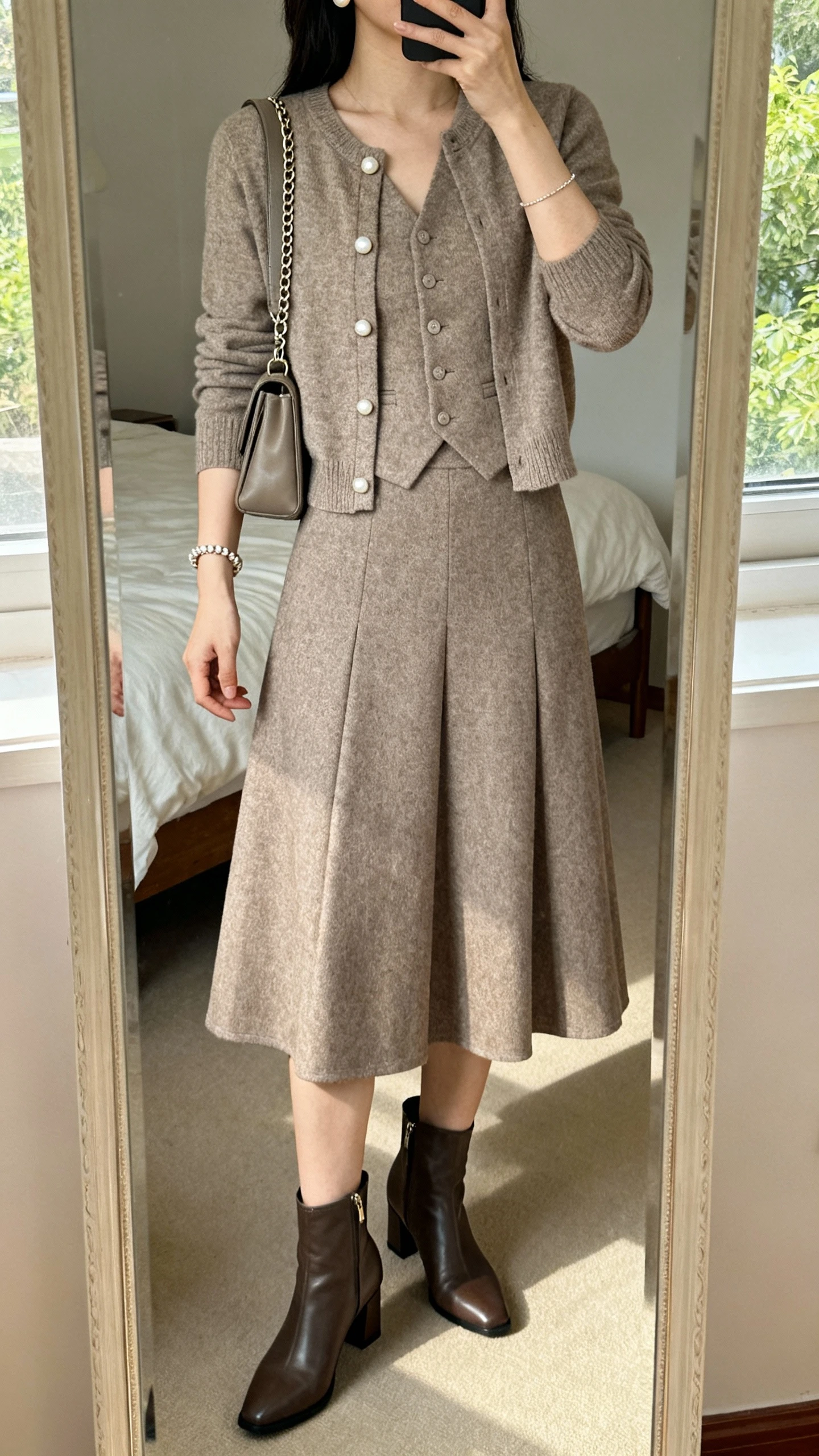 iPhone mirror selfie of a woman in a structured A-line skirt with a matching cardigan set (shell + cardigan), classic low-heel ankle boots, pearl studs, delicate chain bracelet, and a medium shoulder bag, face not visible, cropped at shoulders, bedroom mirror, natural daylight, iPhone photo quality.