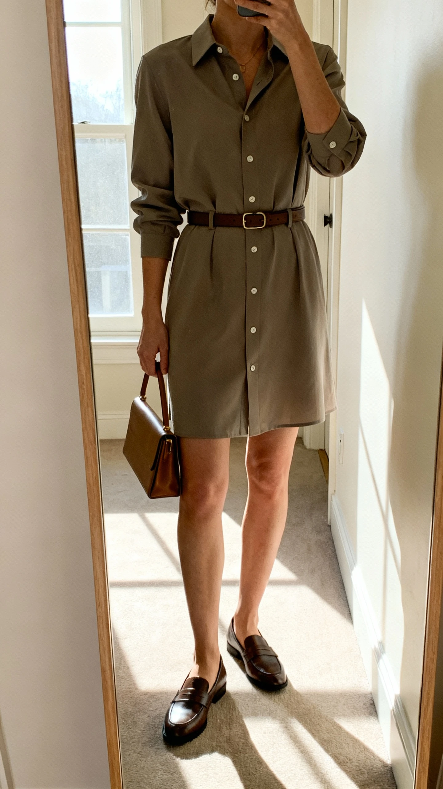 iPhone mirror selfie of a woman in a tailored shirt dress with sleeves, button-front and a belt at the waist, styled with loafers and a slim leather tote, face not visible, cropped at shoulders, hallway mirror, bright window light, iPhone photo quality.