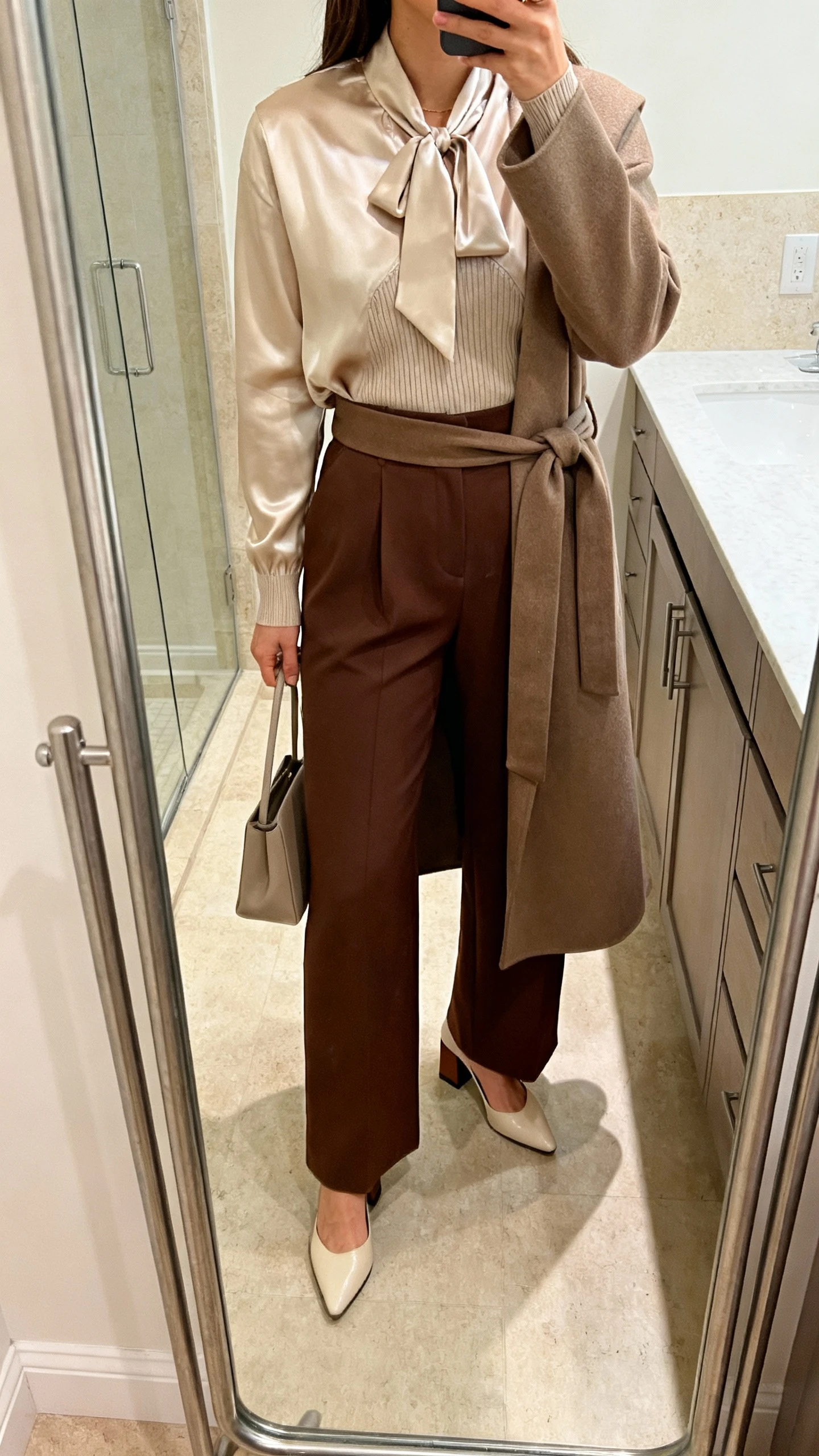 iPhone mirror selfie of a woman in a tie-neck satin blouse layered over a fine knit undershirt, high-waist wide-leg trousers in chocolate, almond-toe block heels, and a tailored wrap coat, holding a minimalist handbag, face not visible, bathroom full-length mirror with soft indoor lighting, iPhone photo quality.