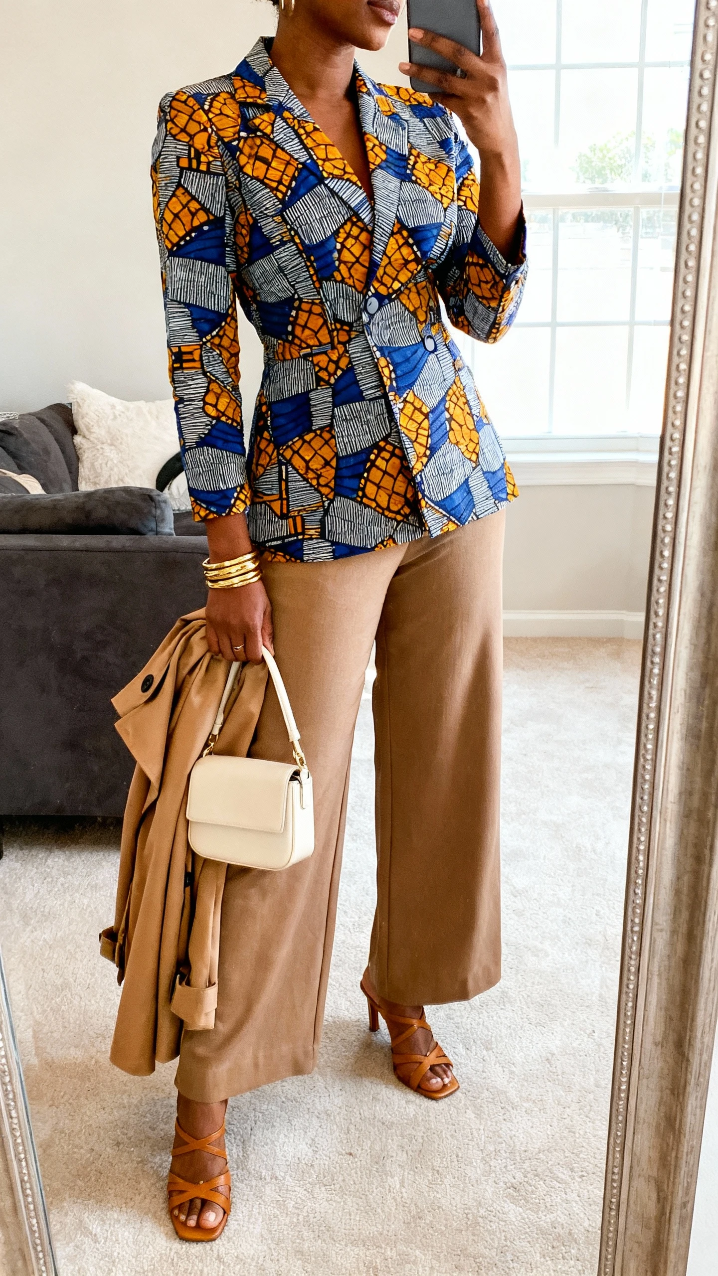 iPhone mirror selfie of a woman in an Ankara print matching set (tailored top and wide-leg trousers) styled with modern neutral accessories: camel trench draped over arm, tan strappy heels, cream mini bag, and gold bangles, face not visible, side angle, living-room mirror, bright window light, iPhone photo quality.