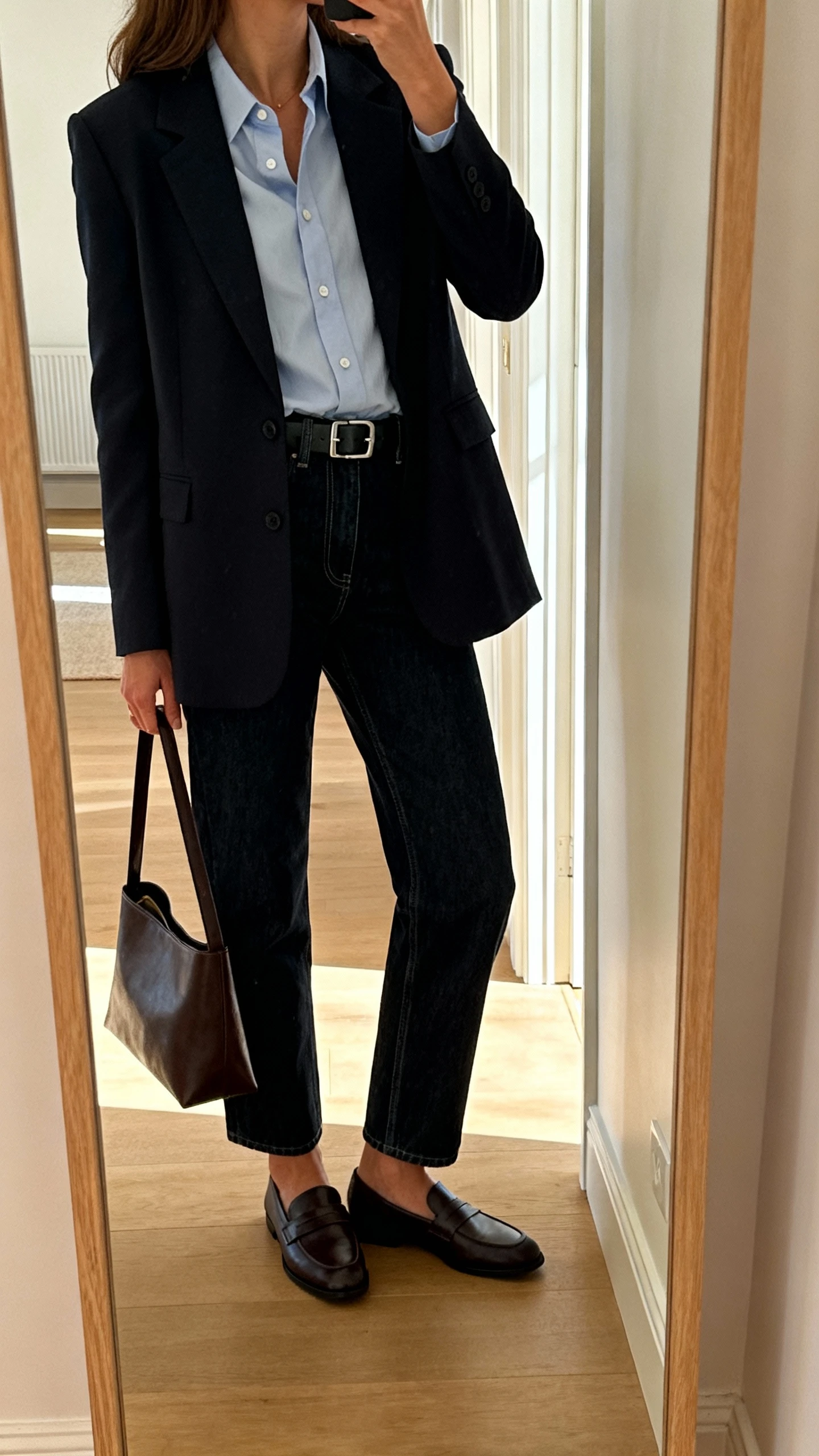 iPhone mirror selfie of a woman in dark-wash straight-leg jeans with a crisp button-up shirt tucked in, tailored blazer, sleek belt, loafers, and a simple leather tote, face not visible, side angle, full-length mirror in hallway, soft afternoon light, iPhone photo quality.