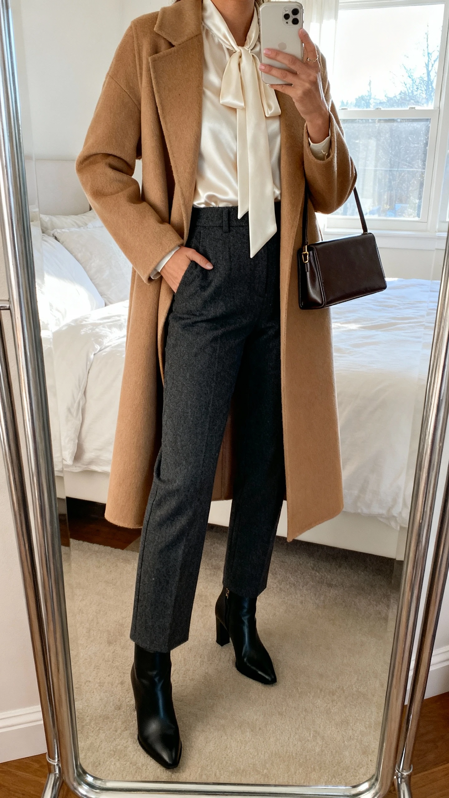 iPhone mirror selfie of a woman in high-waisted charcoal wool-blend straight trousers, ivory satin tie-neck blouse, long camel wool coat, black heeled ankle boots, and a slim leather tote, face not visible, cropped at shoulders in bedroom mirror with natural window light, iPhone photo quality.