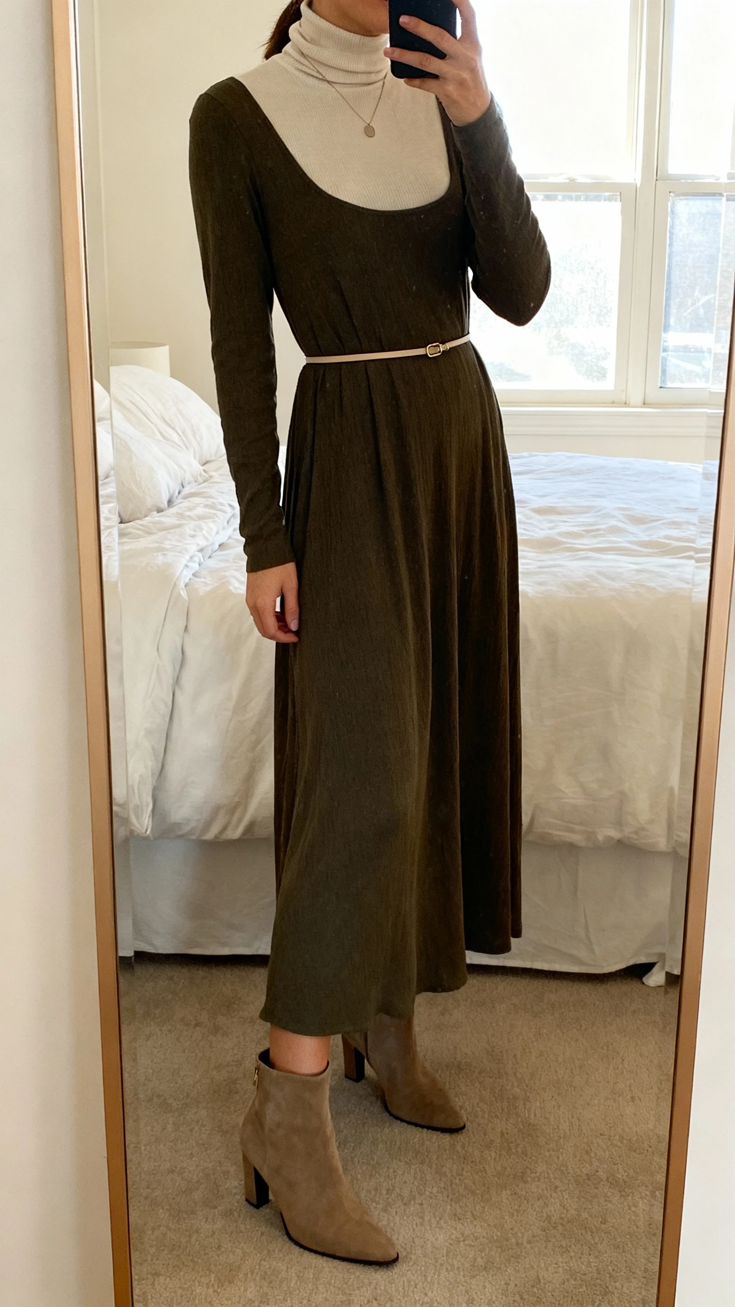 iPhone mirror selfie of a woman layering a fitted turtleneck under a long-sleeve maxi dress, with a thin waist belt, heeled boots, minimal pendant, face not visible, cropped at shoulders, bedroom mirror, soft window light, iPhone photo quality.