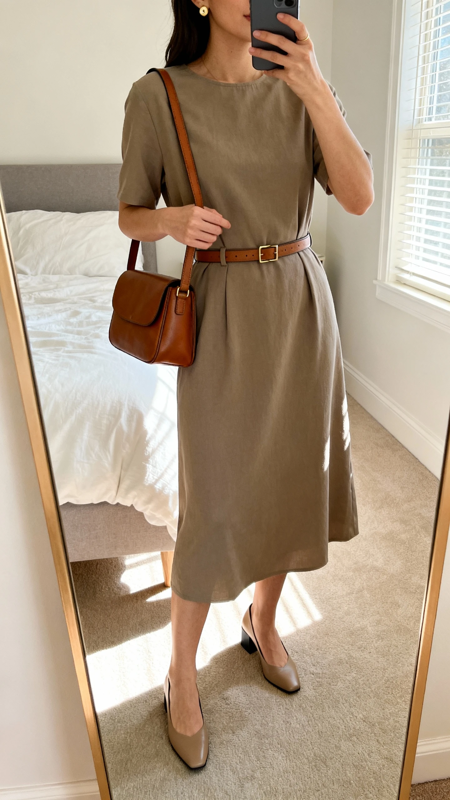 iPhone mirror selfie of a woman wearing a belted midi dress in a solid neutral tone with subtle gold stud earrings, slim leather belt, classic leather shoulder bag, and low block-heel pumps, face not visible, cropped at neck, bedroom mirror with morning window light, iPhone photo quality