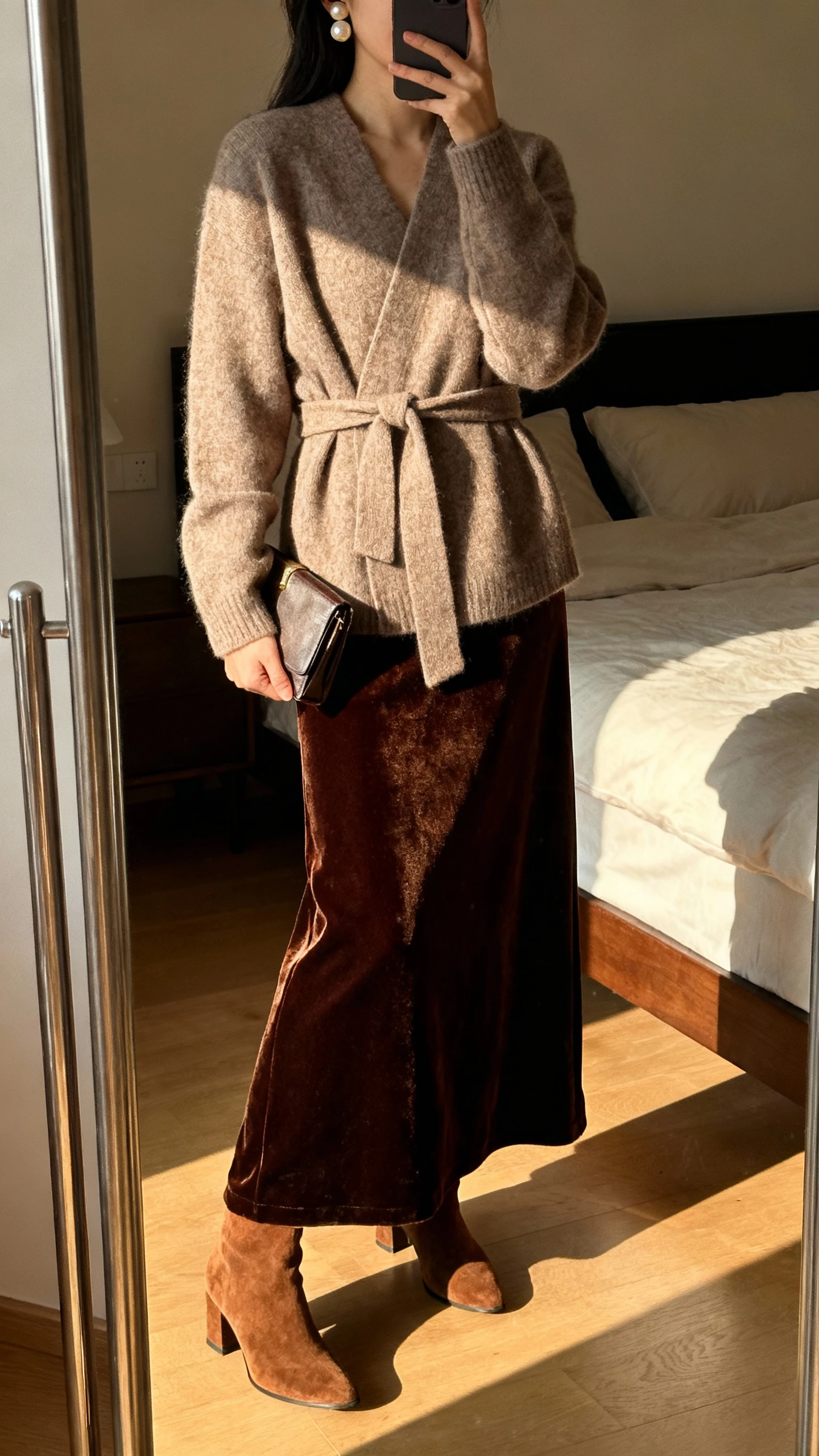 iPhone mirror selfie of a woman wearing a belted wrap sweater over a velvet floor-length maxi skirt with suede heeled boots, pearl studs, and a small clutch tucked under arm, face not visible, cropped at shoulders, bedroom mirror, warm indoor evening light, iPhone photo quality.