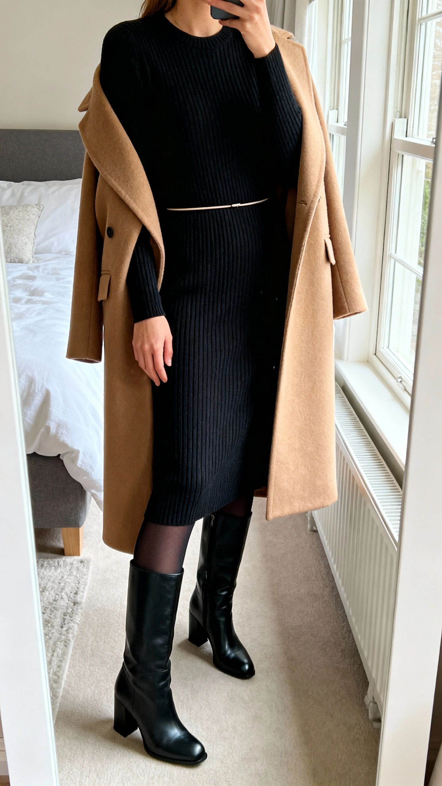 iPhone mirror selfie of a woman wearing a black ribbed knit midi sweater dress with a thin minimal belt, camel tailored wool coat draped over shoulders, black opaque thermal tights, and black leather knee-high block-heel boots; face not visible, cropped at neck, bedroom mirror with natural morning window light, iPhone photo quality.