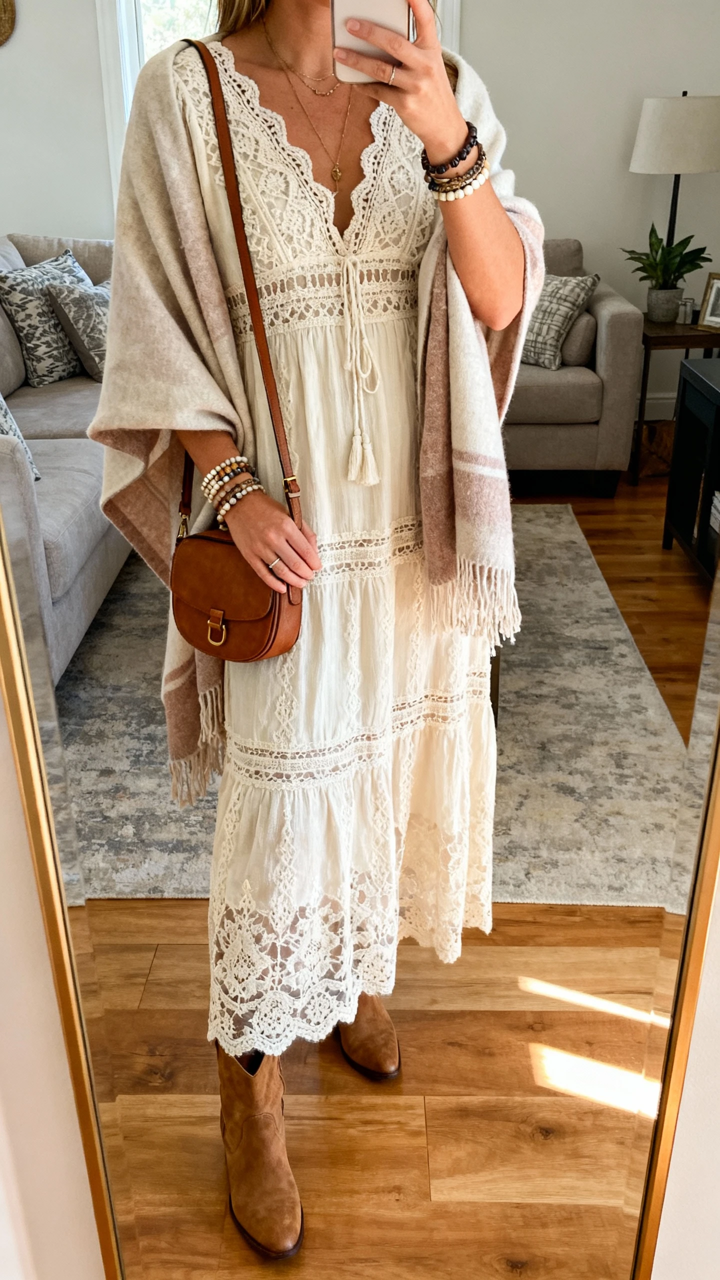 iPhone mirror selfie of a woman wearing a boho lace maxi dress with ankle boots and a soft wrap/shawl, layered bracelets and a small shoulder bag, face not visible, cropped at neck, living room mirror, warm indoor lighting, iPhone photo quality