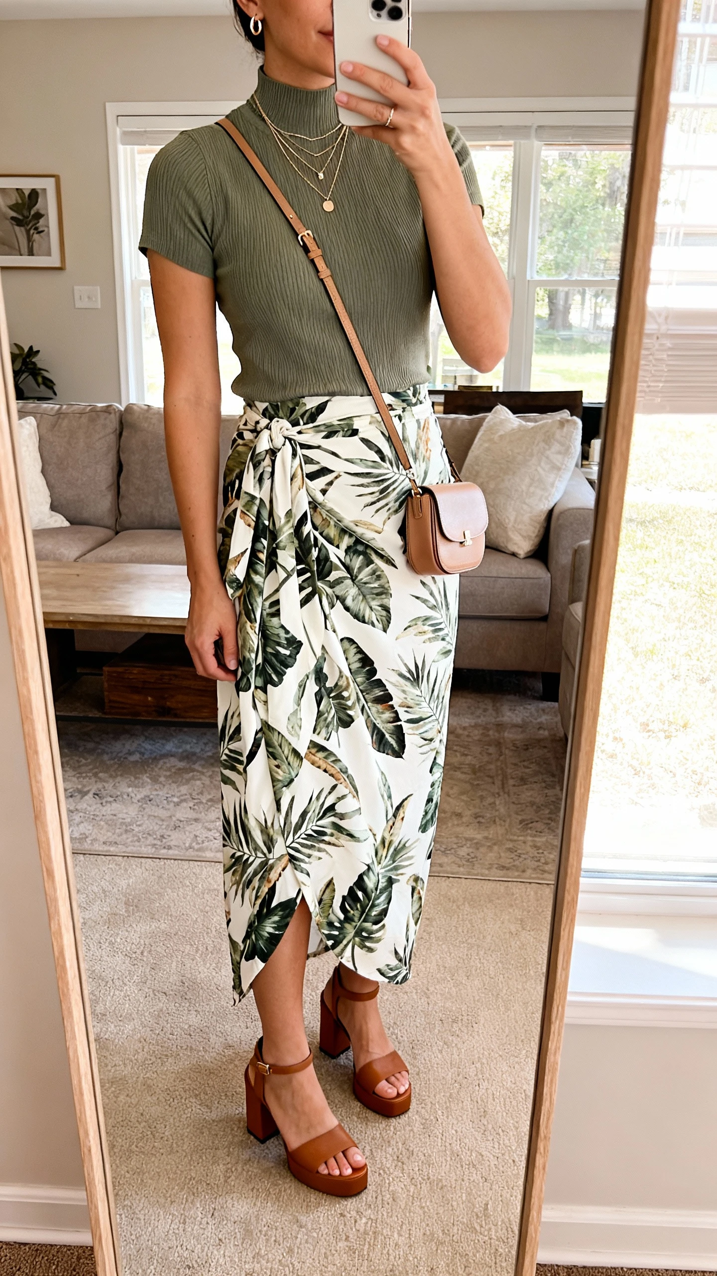 iPhone mirror selfie of a woman wearing a botanical printed wrap maxi skirt, fitted knit tee with a high neckline, block-heel sandals, compact crossbody, layered delicate necklaces, and small hoops, face not visible, cropped at shoulders in a living room full-length mirror, natural window light, iPhone photo quality.