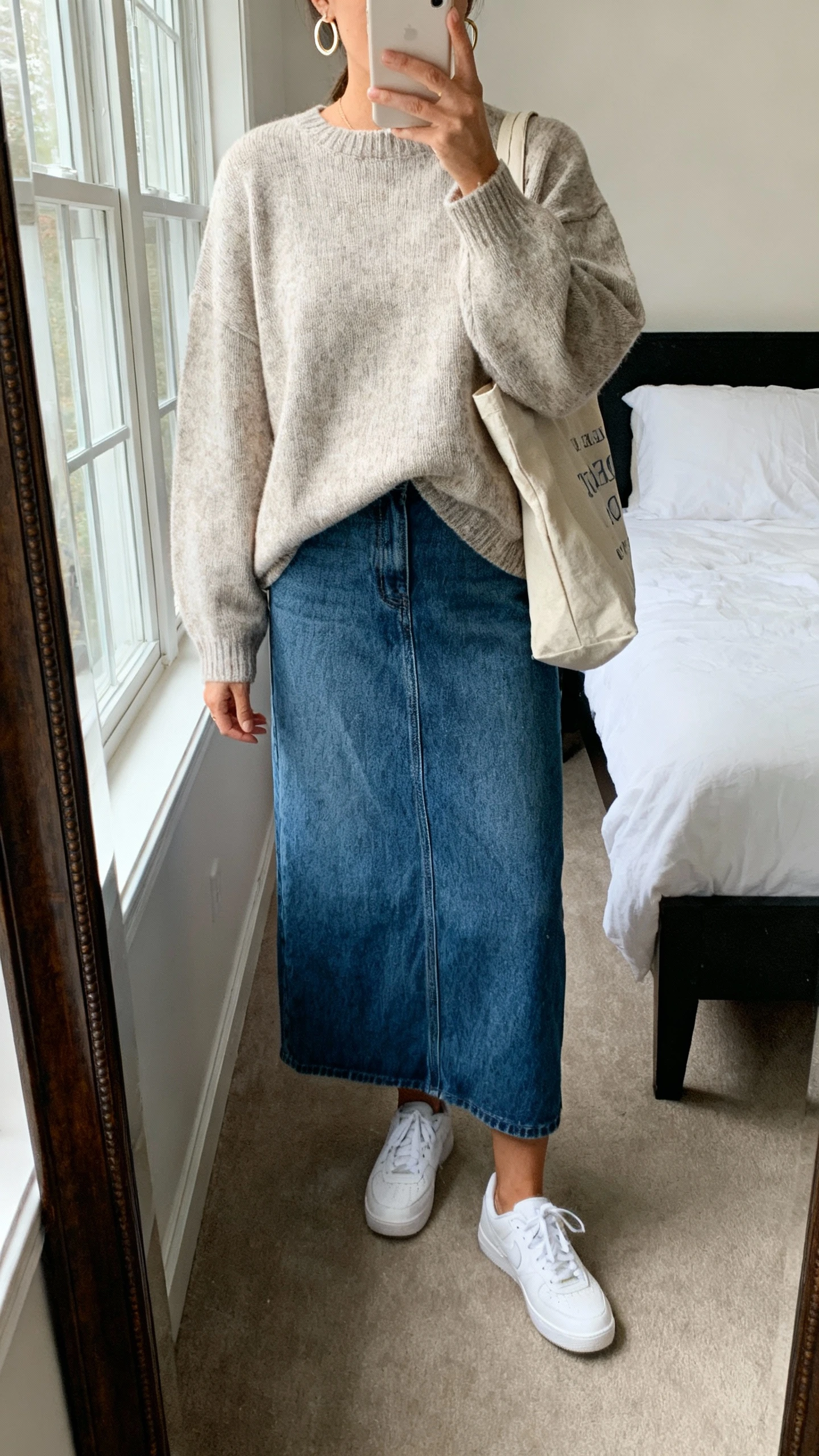 iPhone mirror selfie of a woman wearing a boxy crewneck sweater with a denim maxi skirt and clean white sneakers, simple hoop earrings, and a canvas tote, face not visible, cropped at shoulders, bedroom mirror, natural window light, iPhone photo quality.