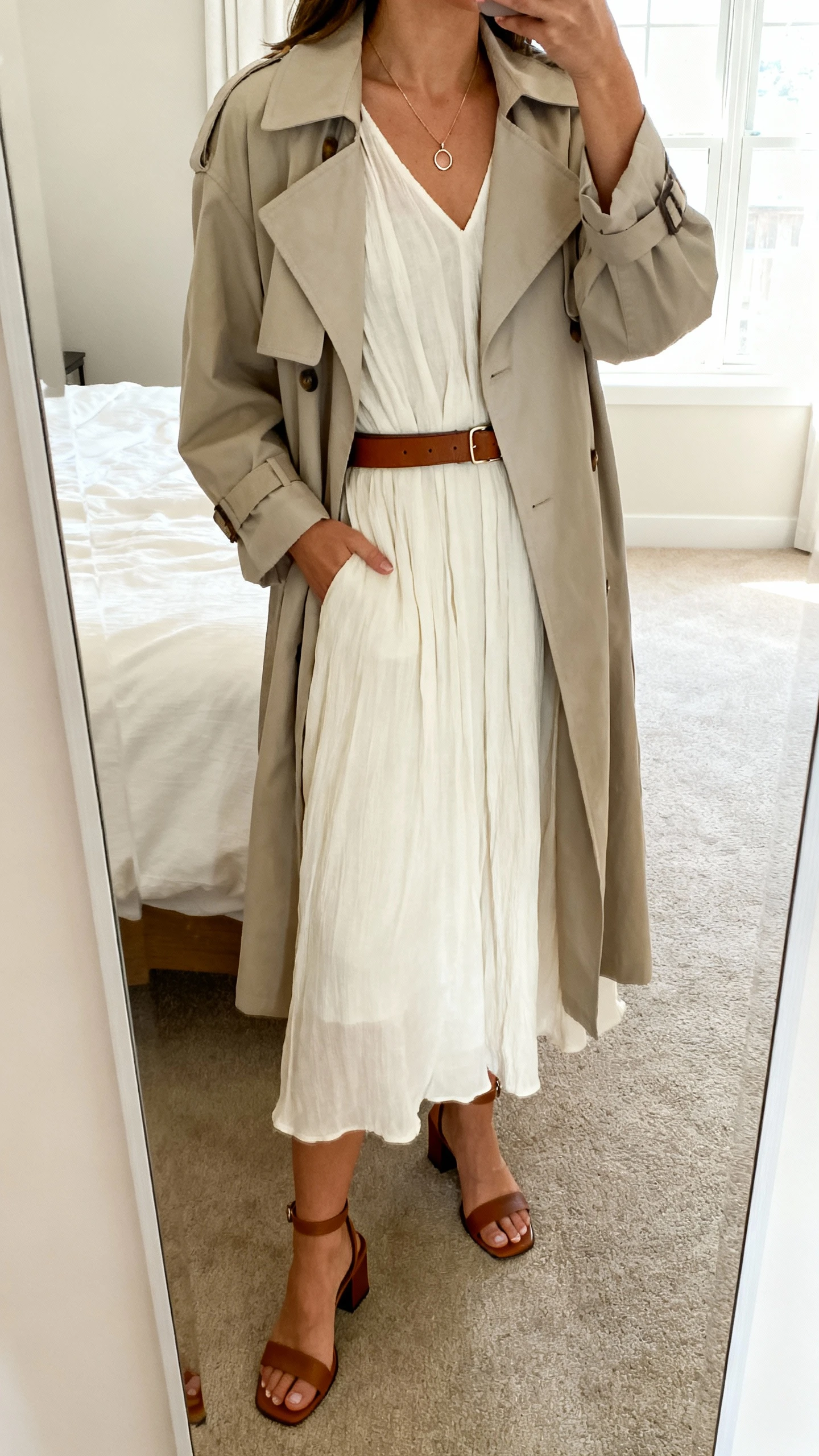 iPhone mirror selfie of a woman wearing a breezy midi dress layered with a lightweight trench, delicate pendant necklace, slim leather belt, and low block-heel sandals, face not visible, cropped at neck, bedroom mirror, soft morning window light, iPhone photo quality.