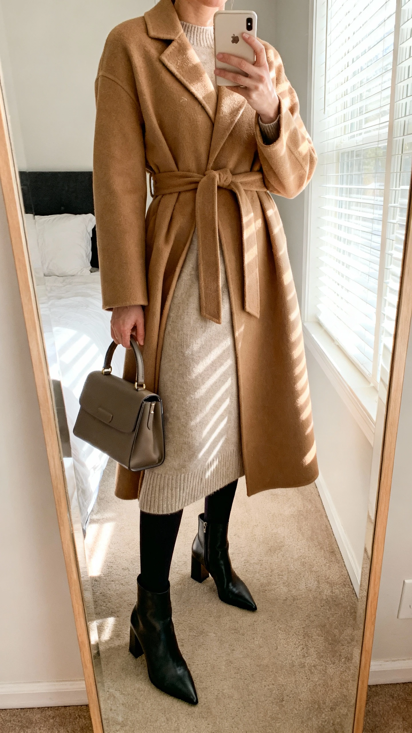 iPhone mirror selfie of a woman wearing a camel wrap coat with belt over a soft midi knit dress (merino/cashmere blend), opaque black thermal tights, pointed-toe block-heel ankle boots, and a structured top-handle bag, face not visible, cropped at neck, bedroom mirror with morning window light, iPhone photo quality.