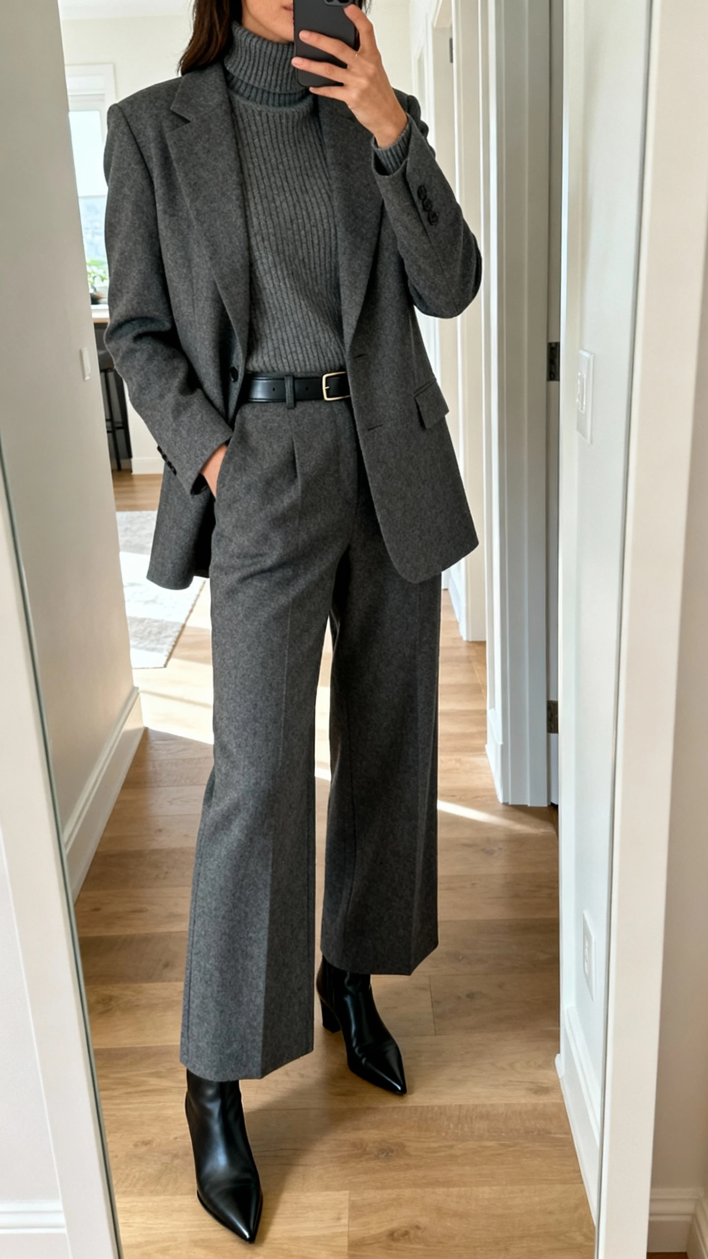iPhone mirror selfie of a woman wearing a charcoal two-piece suit (blazer + wide-leg trousers) with a matching fine-knit turtleneck, sleek black pointed-toe ankle boots, and a minimal leather belt; face not visible, cropped at shoulders, entryway full-length mirror, natural daylight, iPhone photo quality.