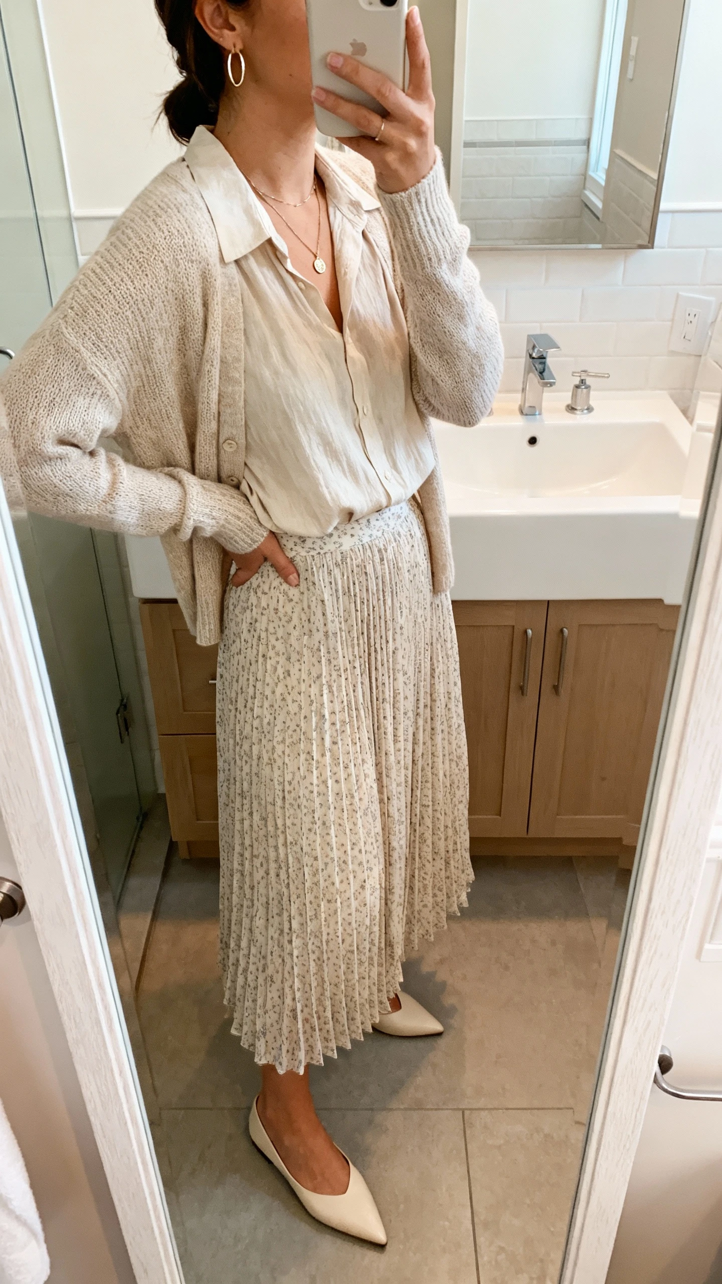 iPhone mirror selfie of a woman wearing a chiffon pleated midi skirt (soft micro-print), a relaxed band-collar blouse, a lightweight knit cardigan, pointed flats, a minimal pendant necklace, and thin hoop earrings; face not visible, side angle in bathroom mirror, indoor soft lighting, iPhone photo quality.
