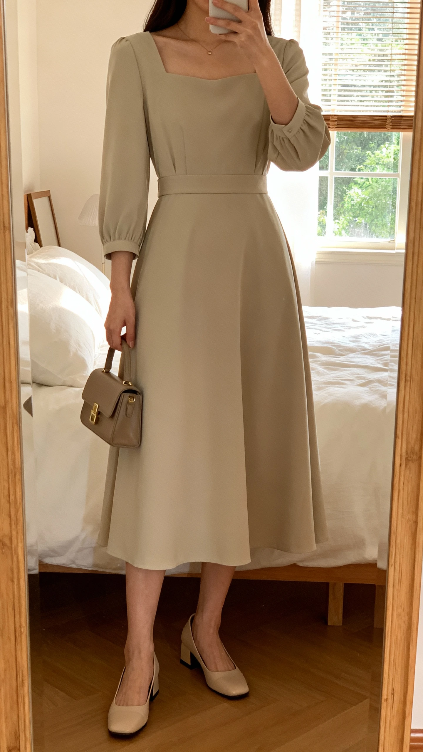 iPhone mirror selfie of a woman wearing a classic A-line midi dress with elbow-length sleeves, defined waist, modest neckline, paired with low block heels and a small structured handbag, face not visible, cropped at neck, bedroom mirror, soft morning window light, iPhone photo quality.