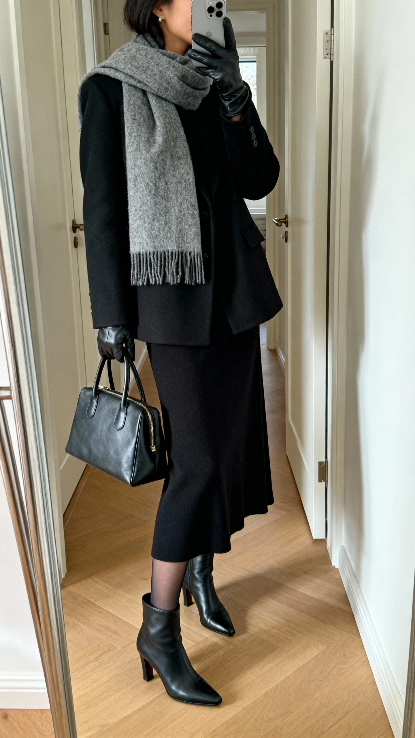 iPhone mirror selfie of a woman wearing a classic black maxi dress styled with winter accessories: wool wrap scarf, tailored black coat, leather gloves, sheer tights, heeled ankle boots, and a structured tote, face not visible, side angle, entryway full-length mirror, natural daylight, iPhone photo quality.